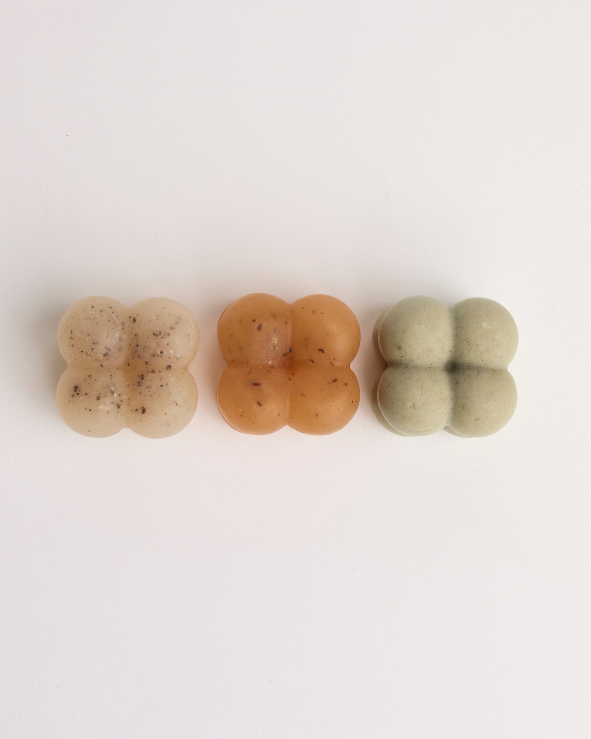 Three soaps shaped like rounded cubes arranged in a row on a white background; colors are beige with specks, amber, and light green.
