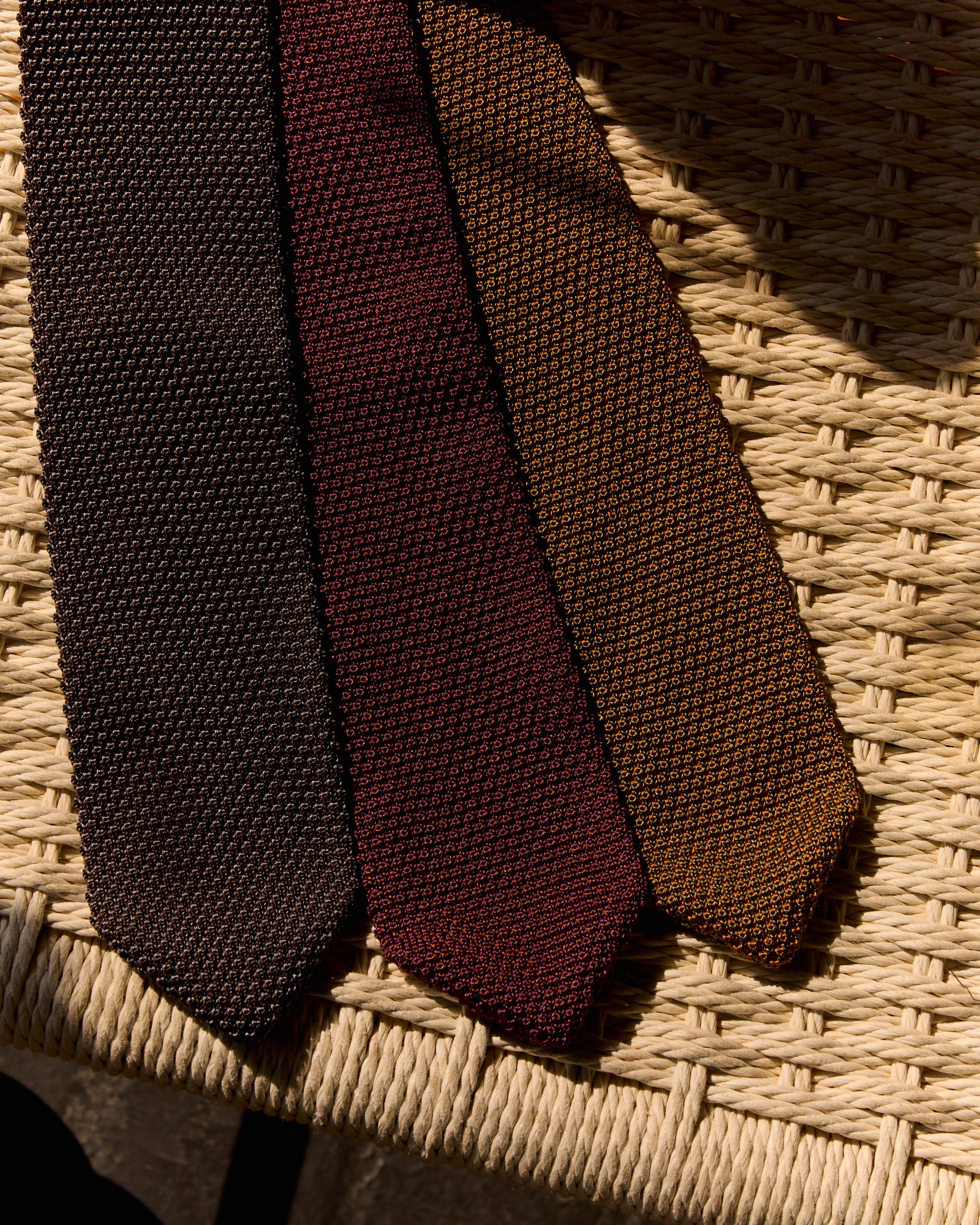 Three textured knit ties in dark brown, burgundy, and ochre are laid side by side on a woven straw chair seat, illuminated by sunlight.