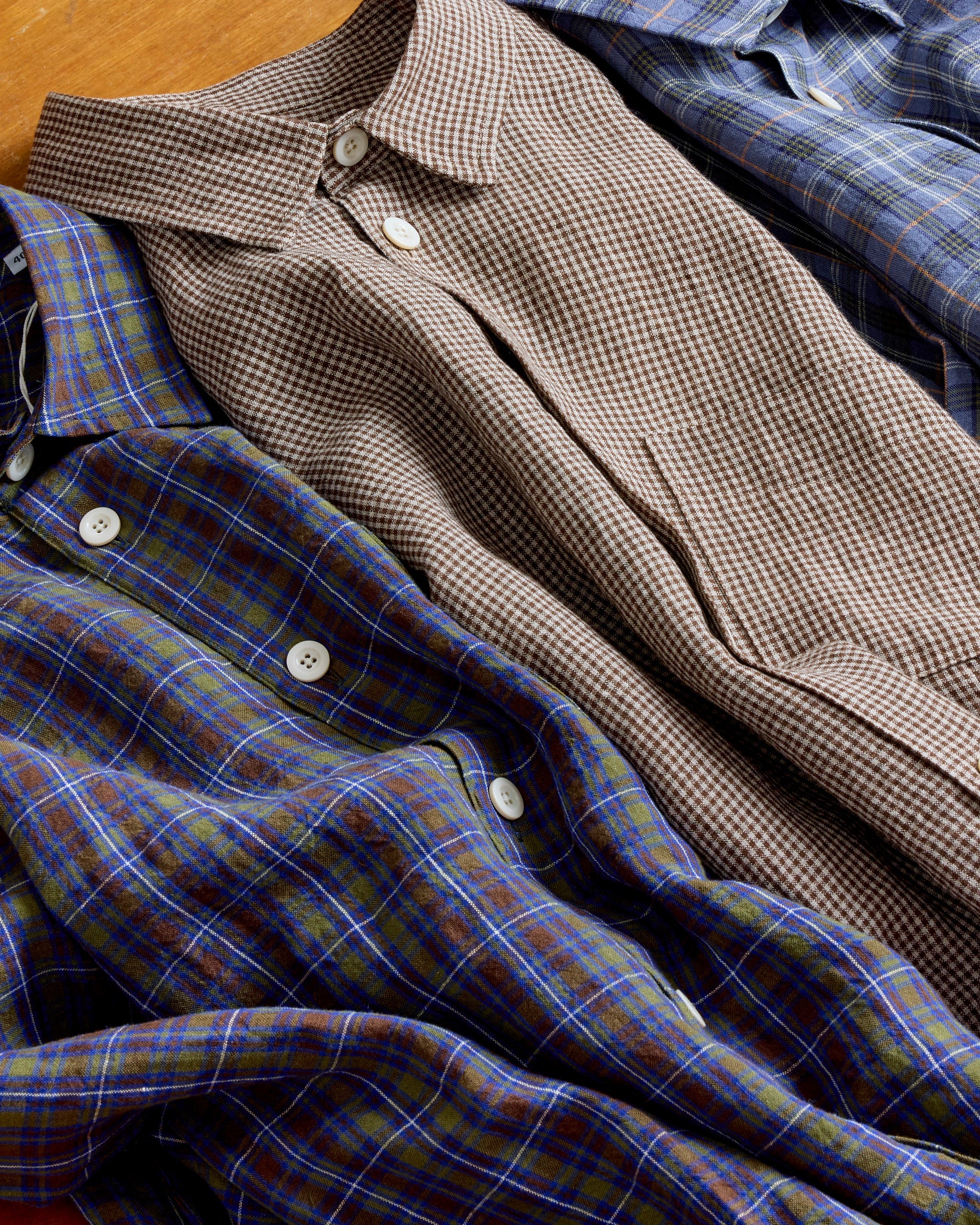 Three neatly folded button-up shirts in plaid and checkered patterns, featuring blue, green, and brown tones, laid side by side on a wooden surface.