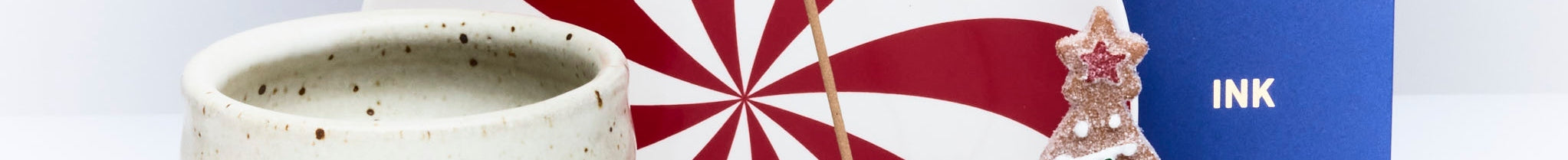 A round speckled ceramic cup, a red and white pinwheel design, a blue book labeled INK, and a Christmas tree-shaped cookie with icing and sprinkles are arranged together.