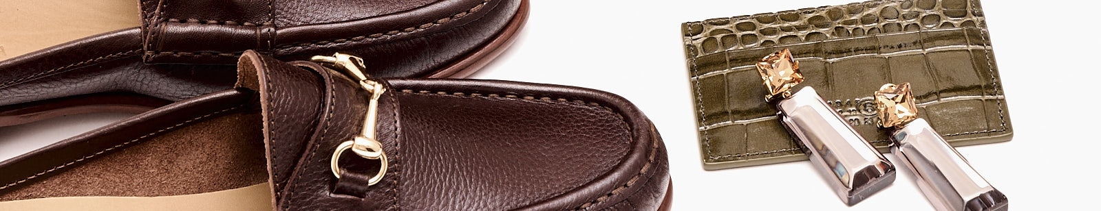 Close-up of brown leather loafers with gold buckle detail, an olive green crocodile-textured wallet, and two tubes of lipstick with gold caps arranged on a white background.