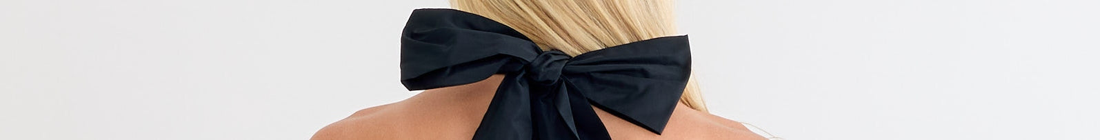 A woman with long blonde hair is seen from behind, wearing a large black bow tied at the back of her neck against a plain white background.