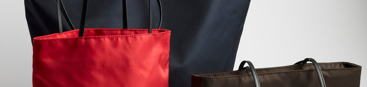Three tote bags are displayed: a red bag in the foreground, a dark brown bag to the right, and a large black bag in the background, all with black handles against a plain background.