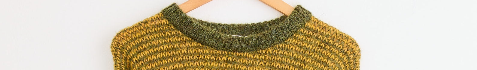 A close-up of a knitted sweater in yellow and green tones, hanging on a wooden hanger against a white background.