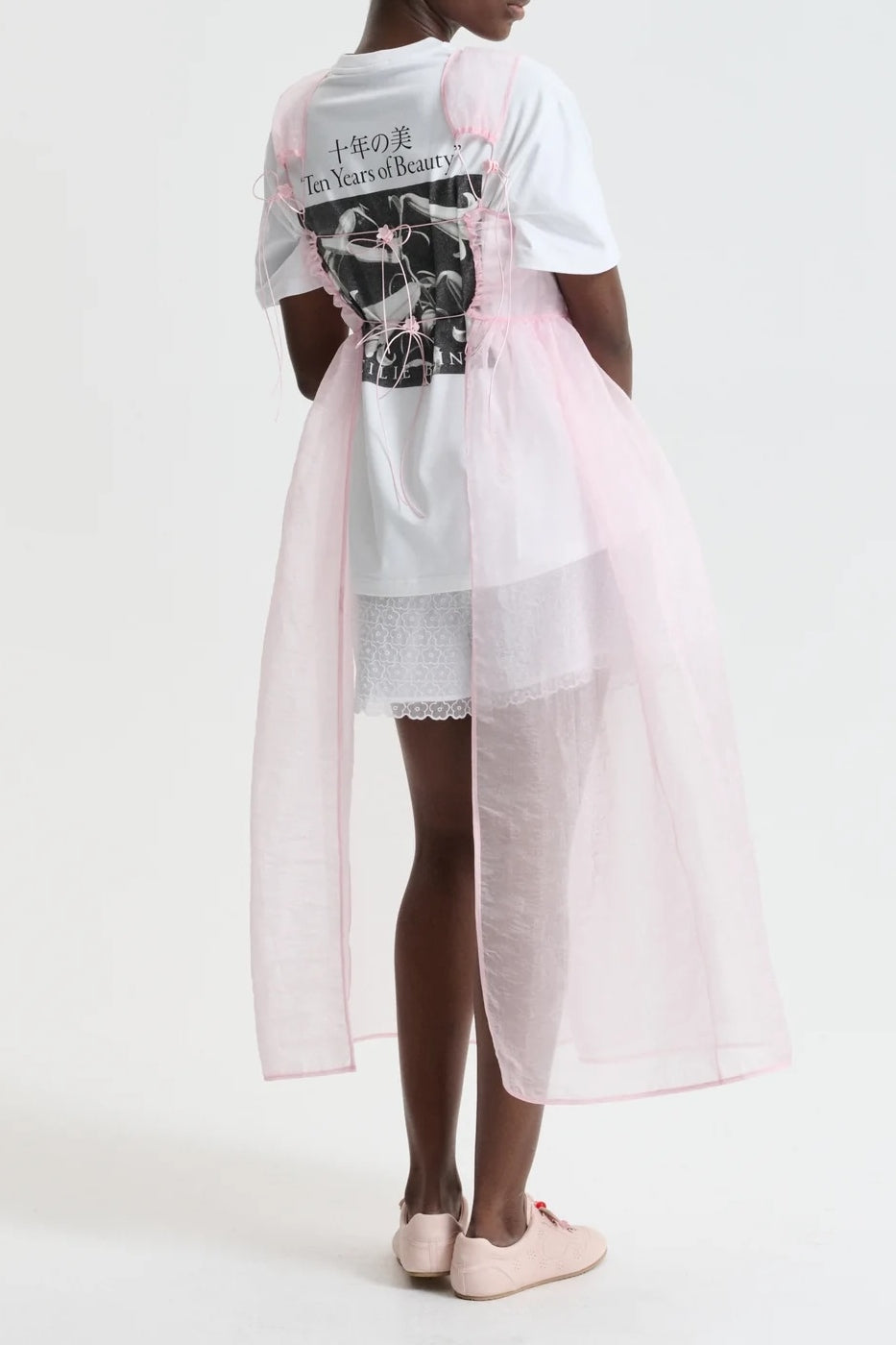 A person stands facing away, wearing a white graphic T-shirt, white shorts with lace trim, and the Cecilie Bahnsen Camilla Overlay Dress in Blush Pink Liquid Myrtia, tied at the back. They wear pale pink shoes. The shirt reads "Ten Years of Beauty" in English and Japanese.