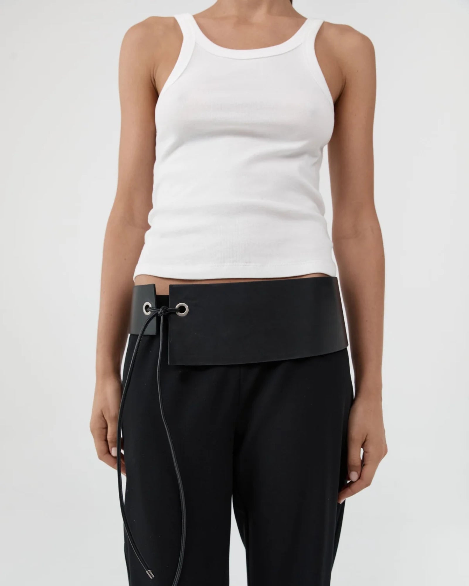 A person wearing a white tank top, black pants, and the ST. AGNI Asymmetric Belt in Black stands against a plain light background.
