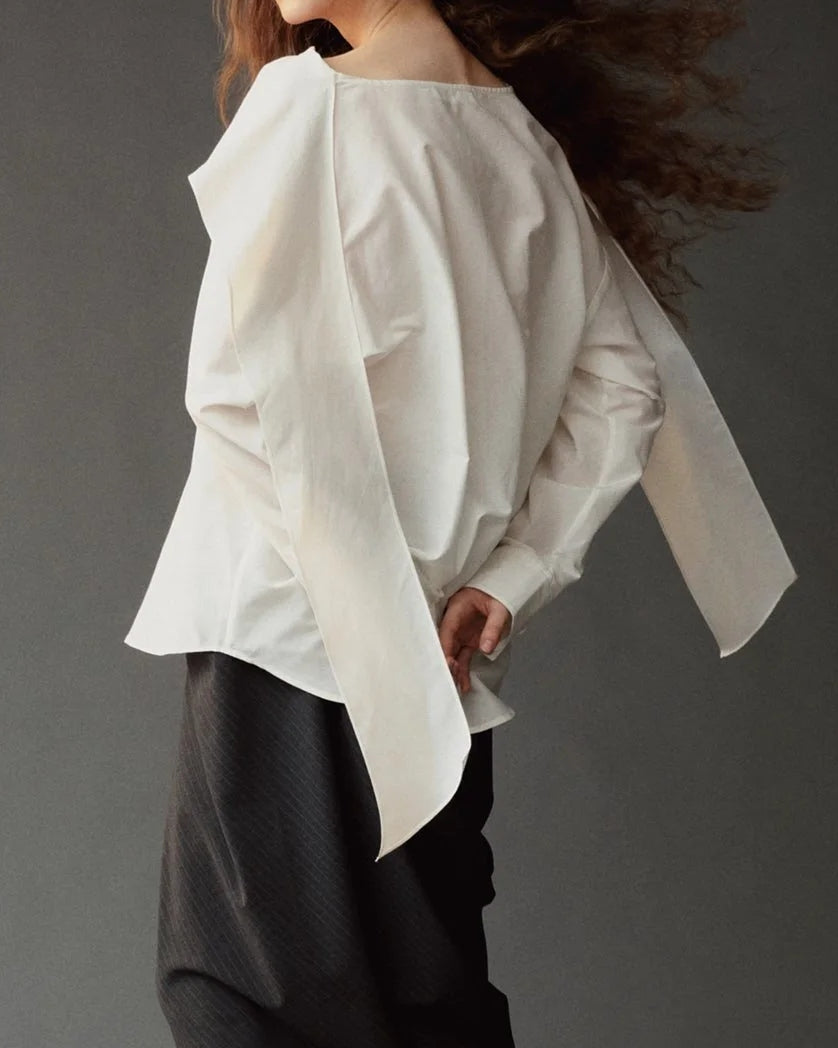 A person with long curly hair stands sideways against a plain grey background, hands behind their back, wearing the Mijeong Park Silk Blend Tie Neck Blouse in Ivory—an oversized fit with wide sleeves—paired with a black skirt.