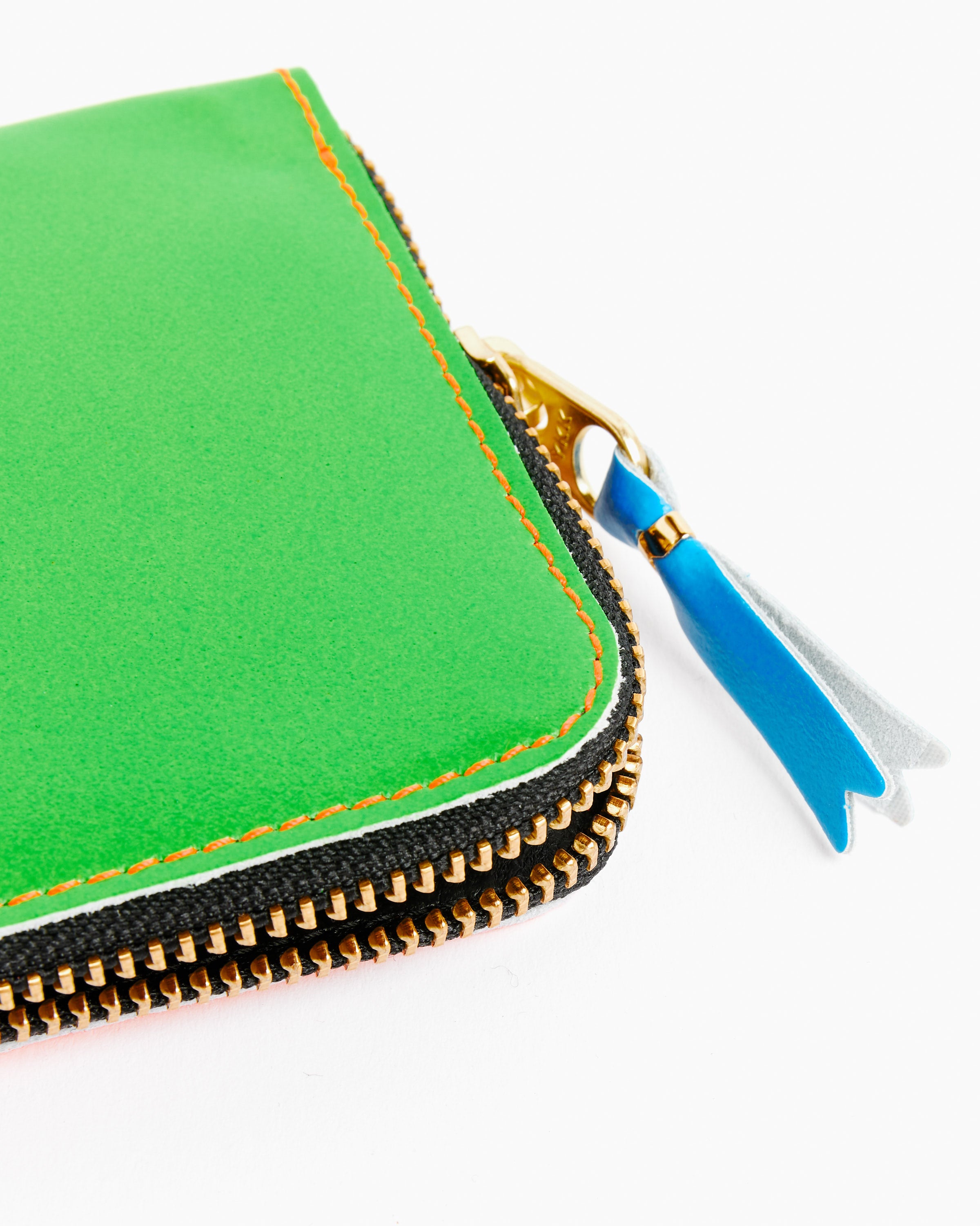 A close-up of the Comme des Garcons Wallet Super Fluo Half Zip Wallet in Green/Orange—green goat leather with orange stitching, gold zipper, blue and silver leather pulls—made in Spain, set against a white background.