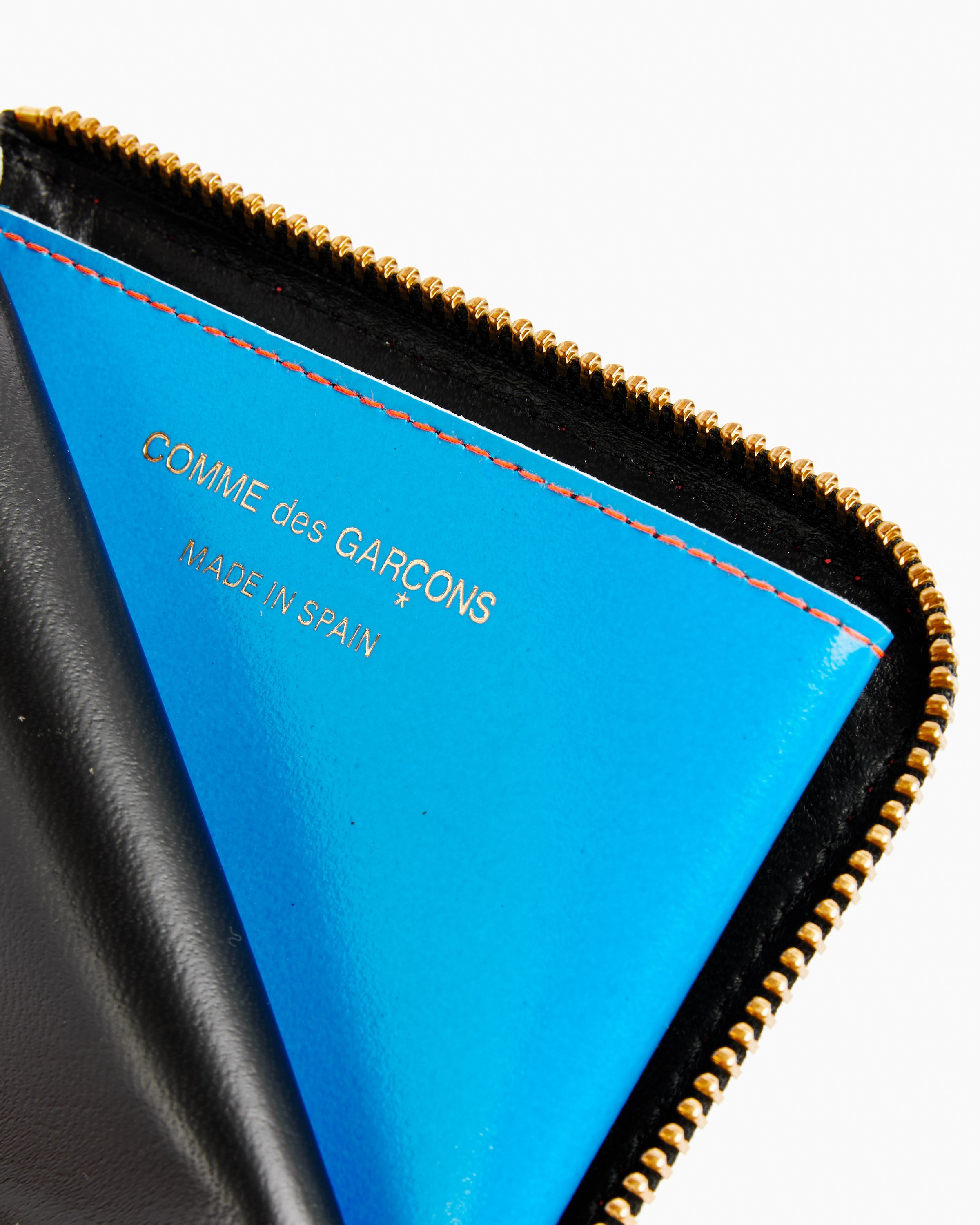Close-up of the Super Fluo Half Zip Wallet in Green/Orange by Comme des Garcons Wallet, partially open to show a bright blue interior with gold COMME des GARÇONS text and made in Spain, plus a gold zipper with black trim.