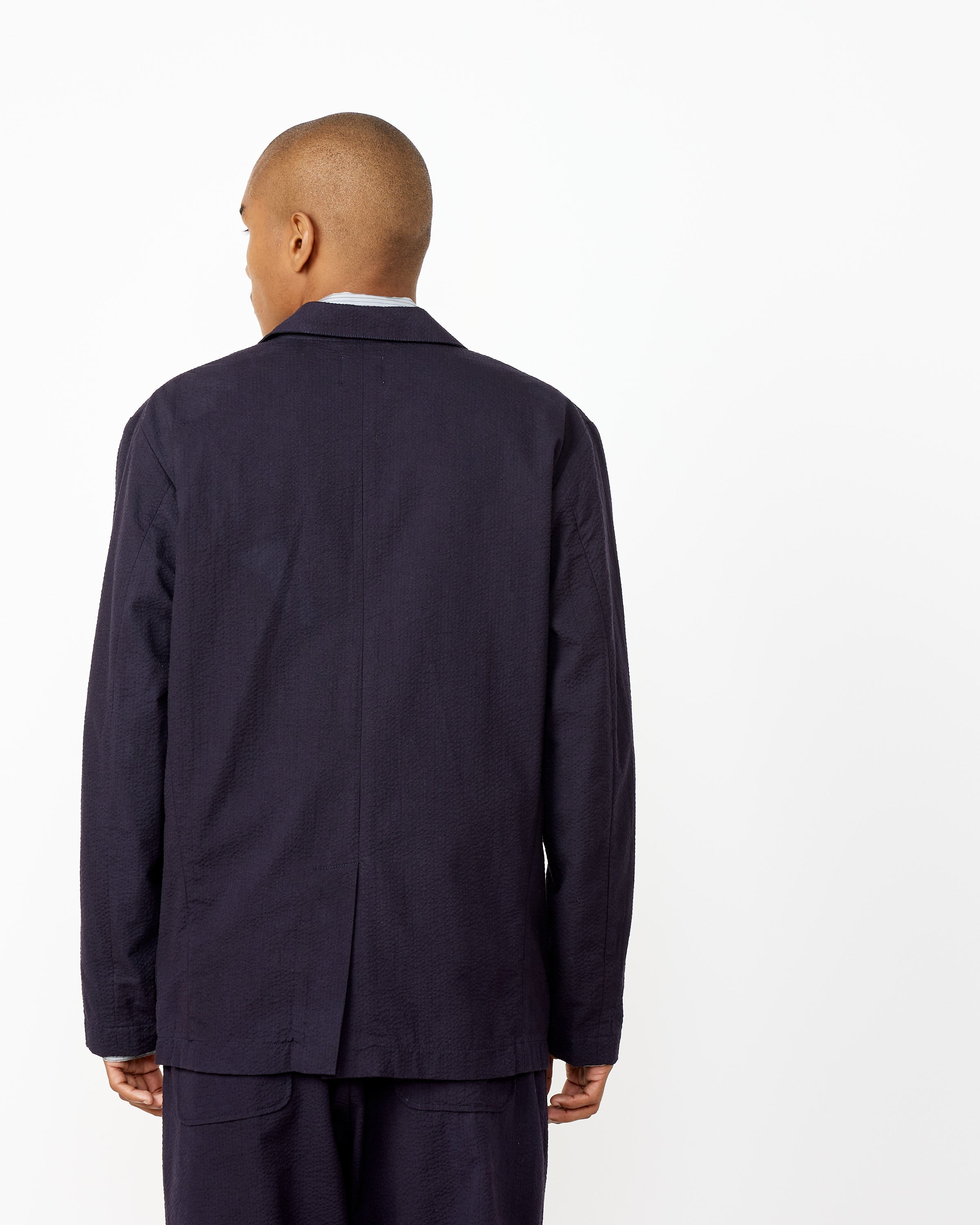 A person with a shaved head, seen from behind, wears the SMOCK Blazer in Seersucker Midnight with matching trousers against a plain white background.