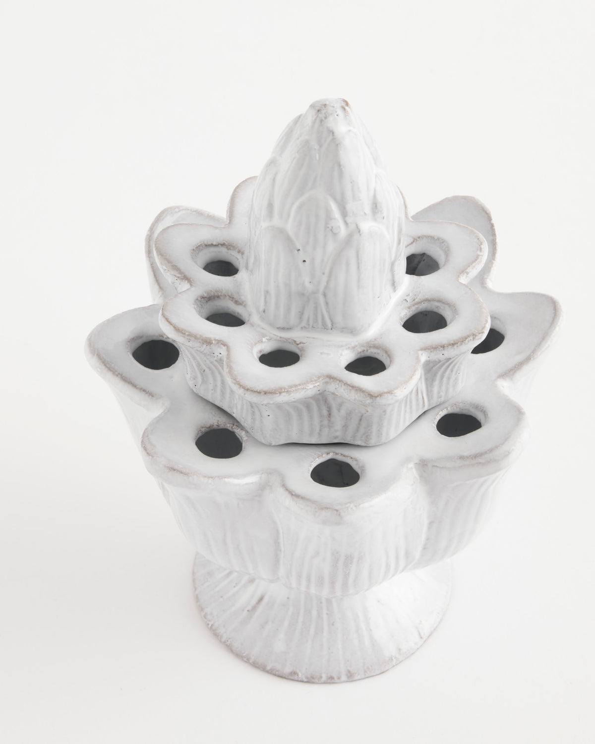 The Pique Flower Holder by Astier de Villatte is a white ceramic piece handmade in Paris, characterized by flower-like layers, circular holes, and a pointed top reminiscent of an artichoke or lotus. Its smooth surface features subtle brown accents and is photographed against a plain white background.