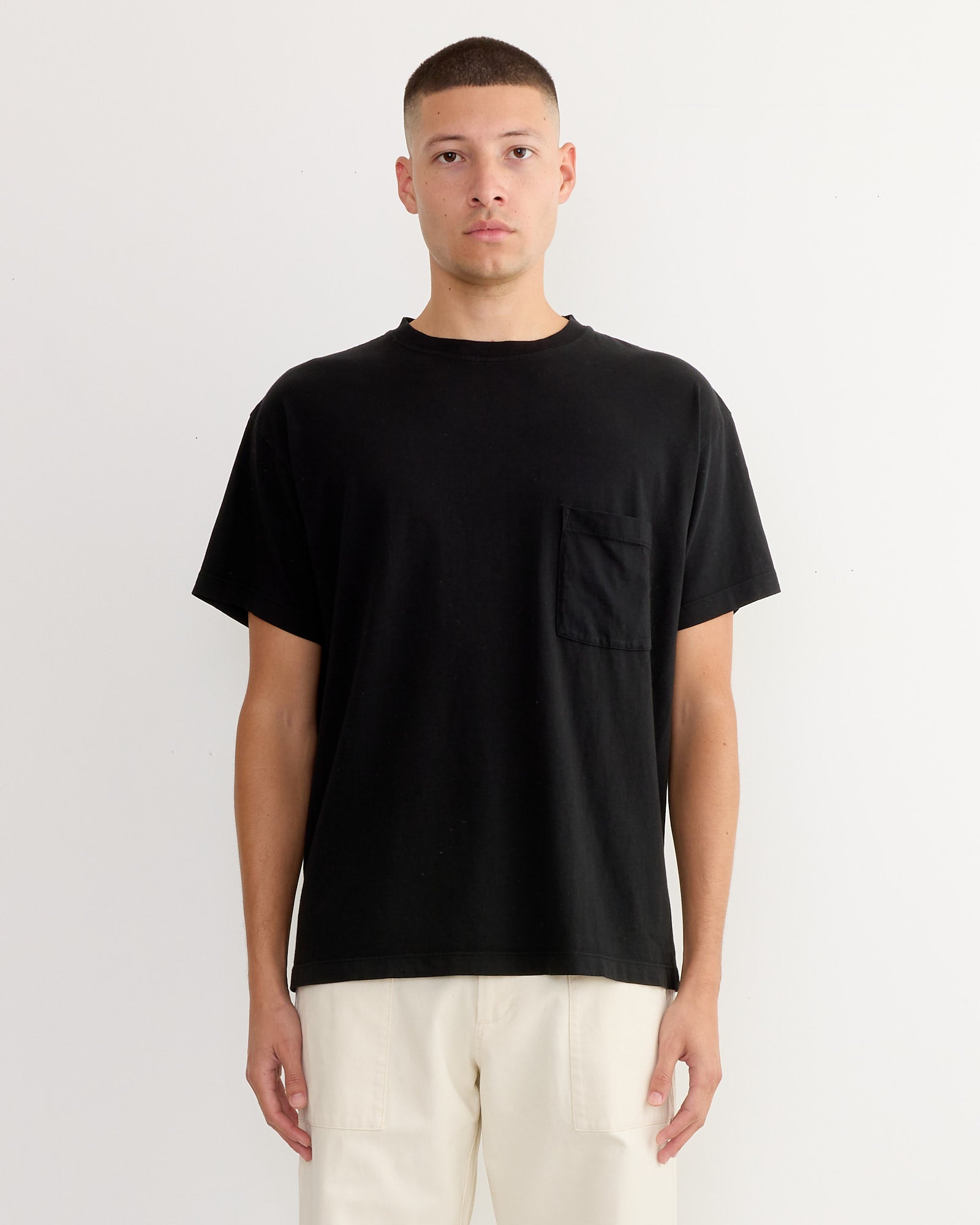A person wears a loose black SMOCK Big Pocket Tee from the 3 Pack, made of soft combed cotton jersey, with light beige pants, standing facing forward against a plain white background.