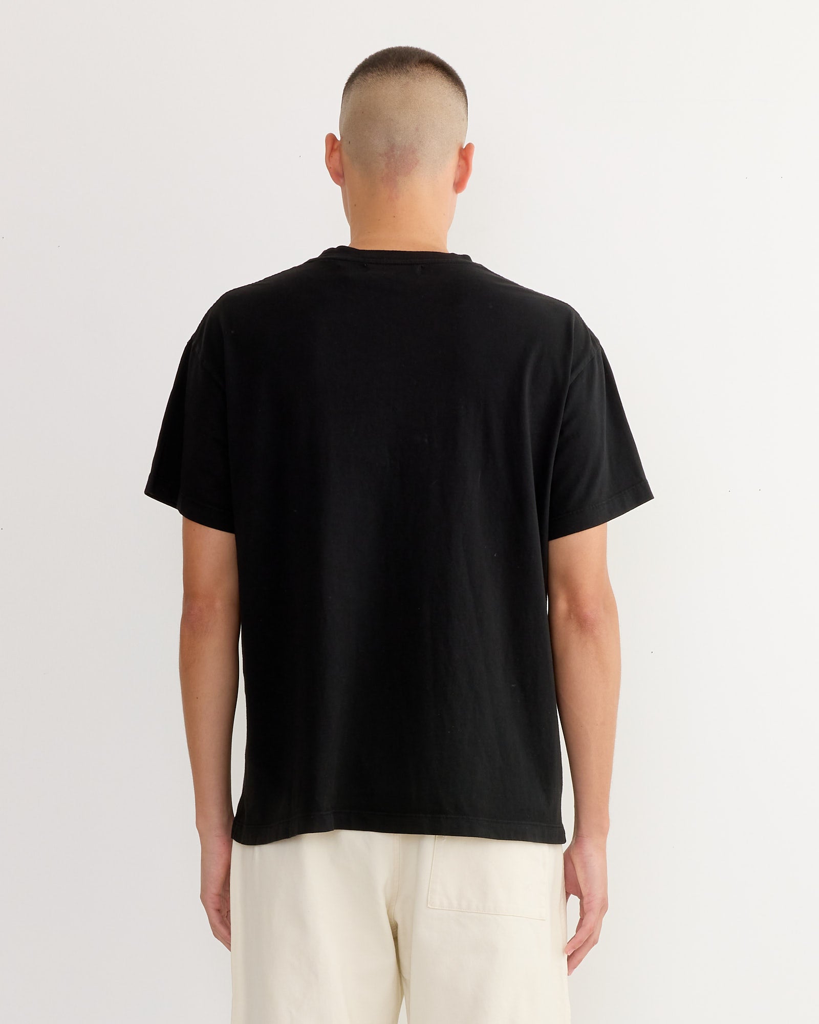 A person with a buzz cut stands facing away from the camera, wearing the SMOCK Big Pocket Tee 3 Pack in Black and light-colored pants against a plain white background.