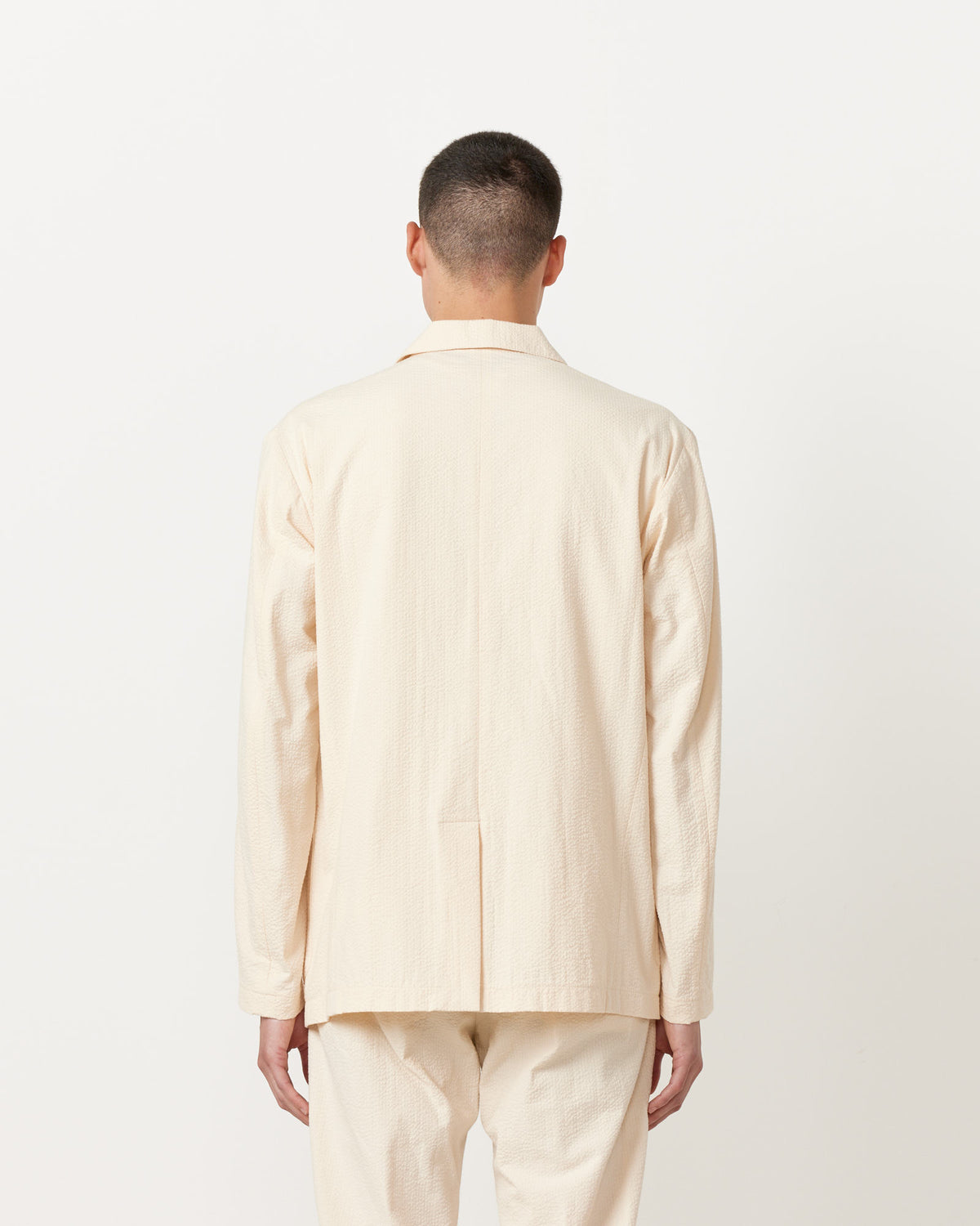 A person with short dark hair, seen from behind, wears the SMOCK Seersucker Blazer in Cream with matching pants, standing against a plain white background.