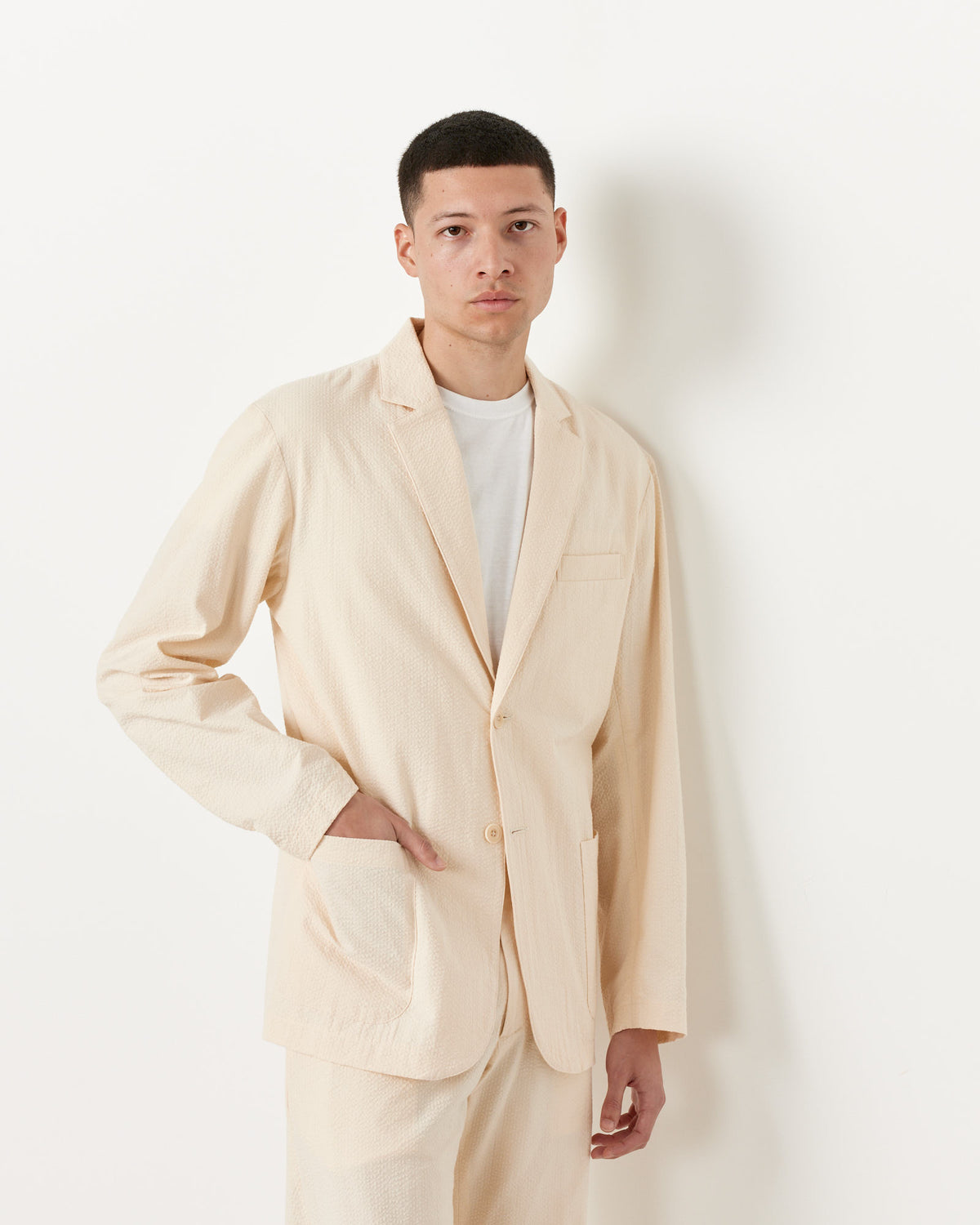 A man wears the SMOCK Seersucker Blazer in Cream with matching pants, standing against a white background with one hand in his pocket and a neutral expression.