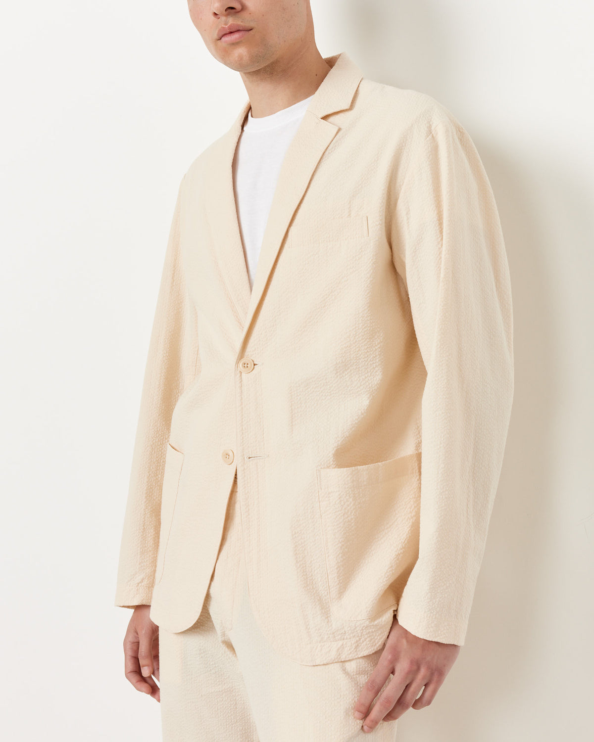 A person wears the SMOCK Seersucker Blazer in Cream—crafted from textured Japanese cotton with two buttons and large front pockets—over a white tee and matching pants, standing against a plain white background.