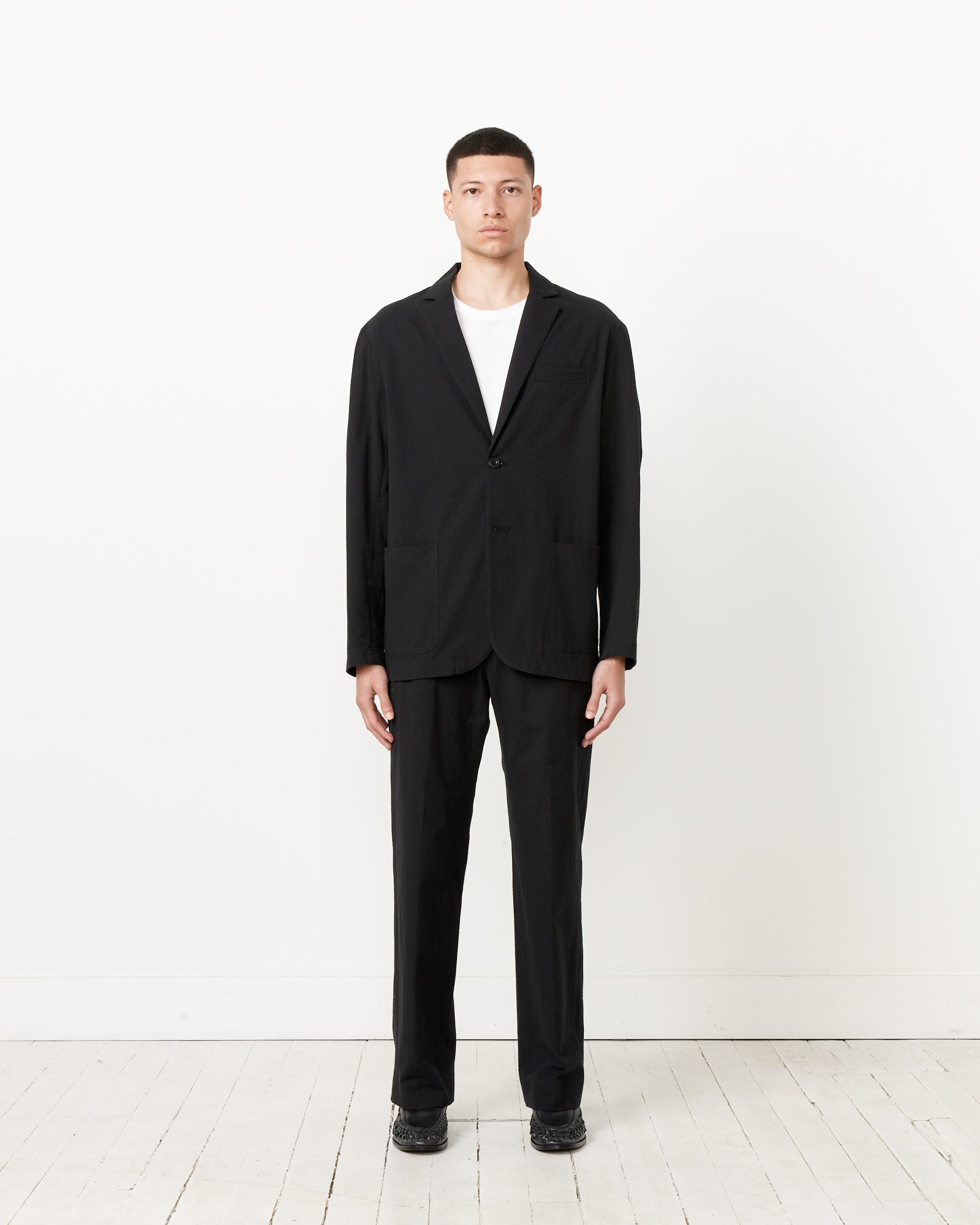 A person stands facing forward against a plain white wall, wearing the SMOCK Seersucker Blazer in Black over a white t-shirt with black shoes, on a white wooden floor.