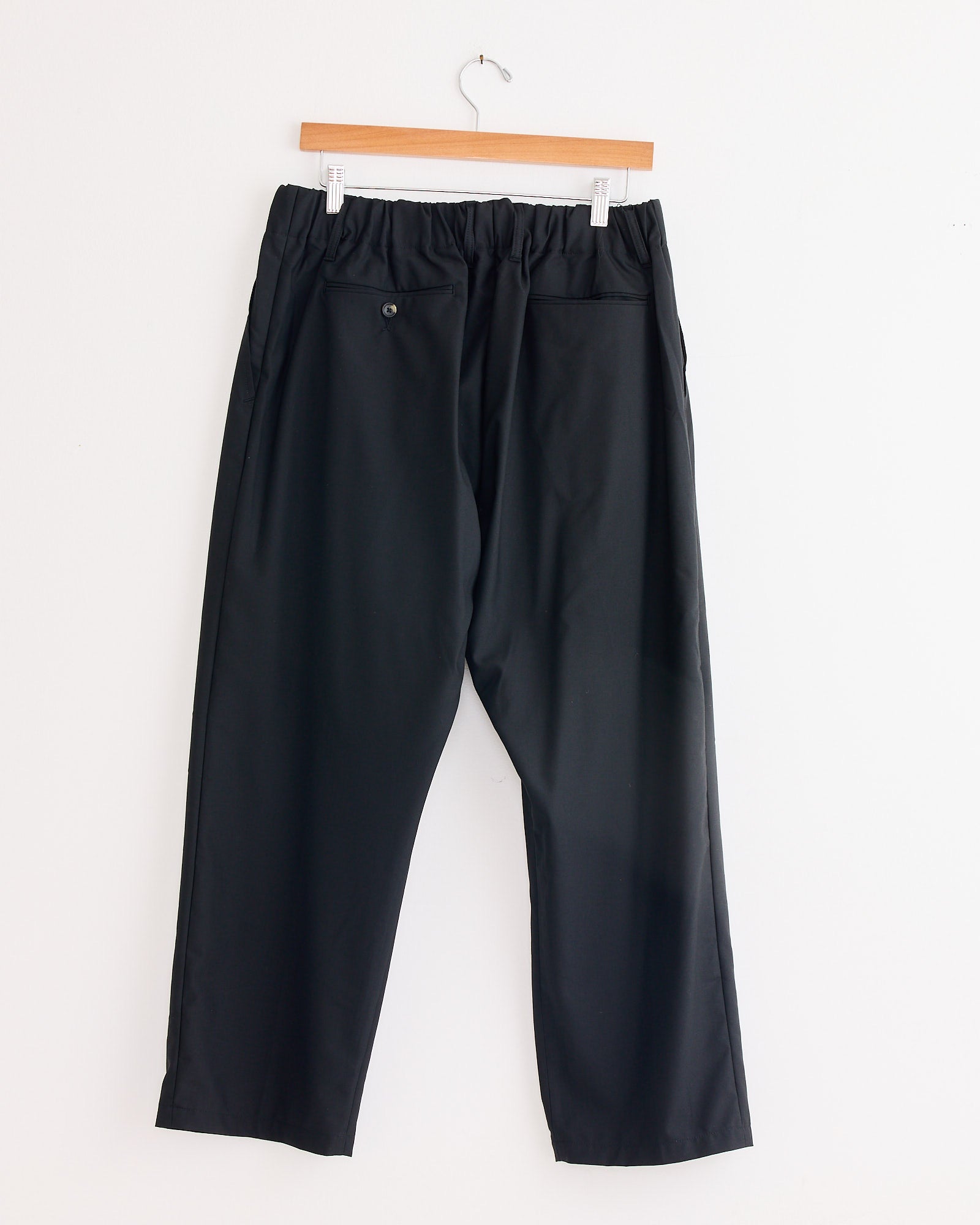 The Sillage Baggy Trouser in Black, featuring a cropped cut, elastic waistband, and two back pockets (one with a button), hangs on a wooden hanger against a white wall. Made in Japan.