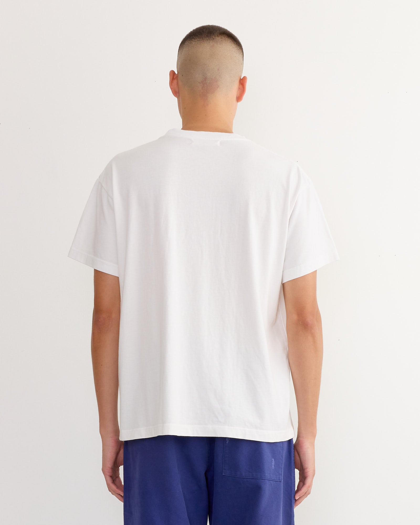 A person with a buzz cut, seen from behind, wears the SMOCK Big Pocket Tee in White and blue pants, standing against a white background.