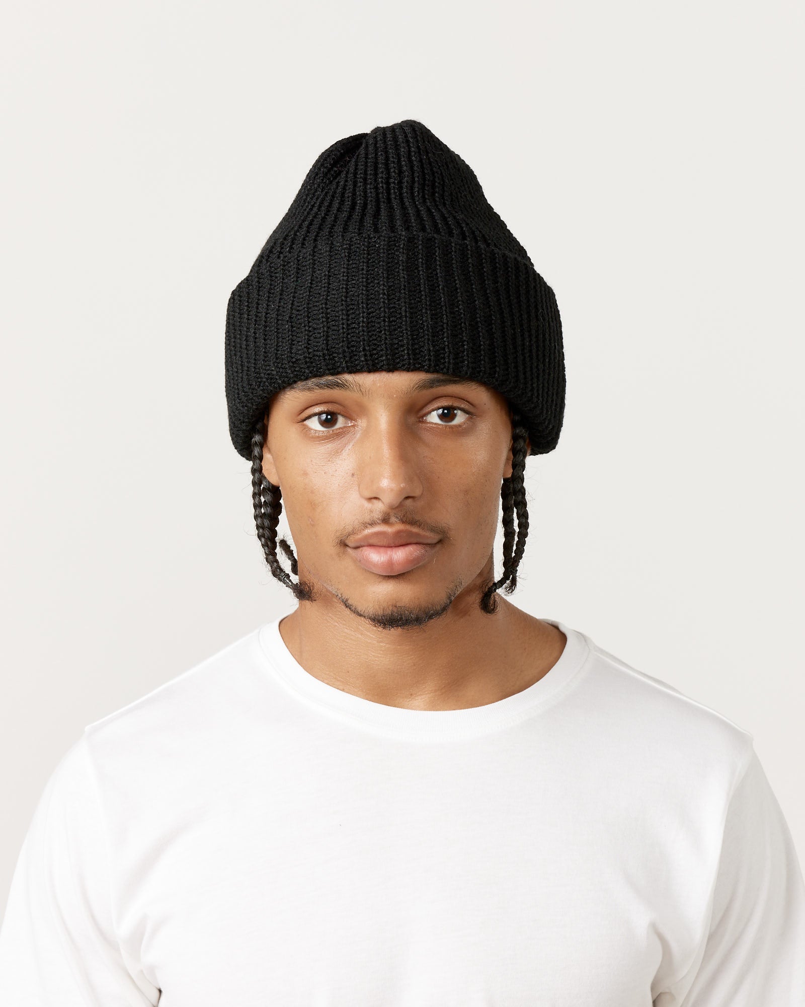 A person with braided hair wears the Rototo 5075 Chunky Beanie in Black and a plain white t-shirt, looking directly at the camera against a light, neutral background.