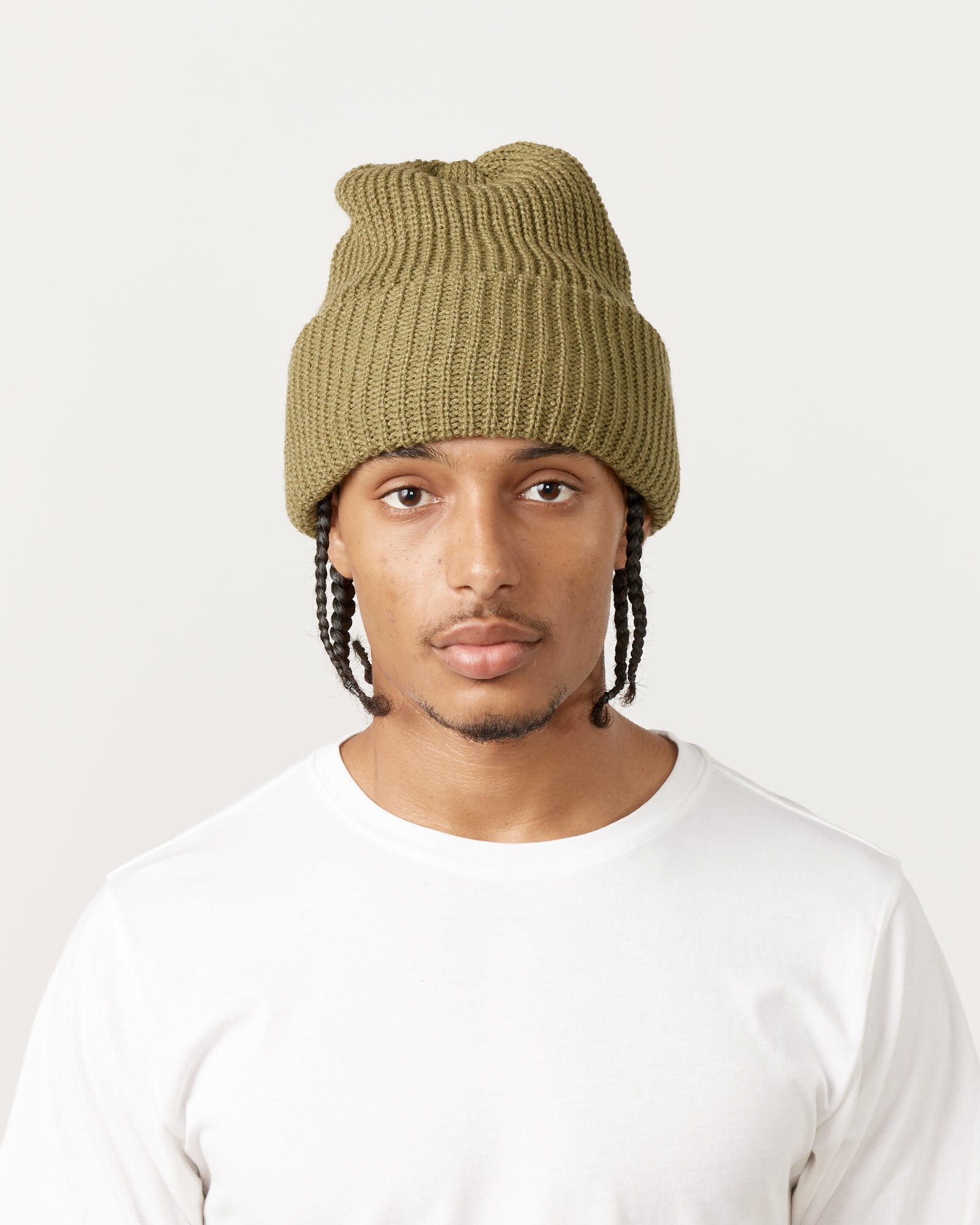 A person wears the Rototo 5075 Chunky Beanie in Light Olive and a white t-shirt, staring neutrally at the camera against a plain light background.