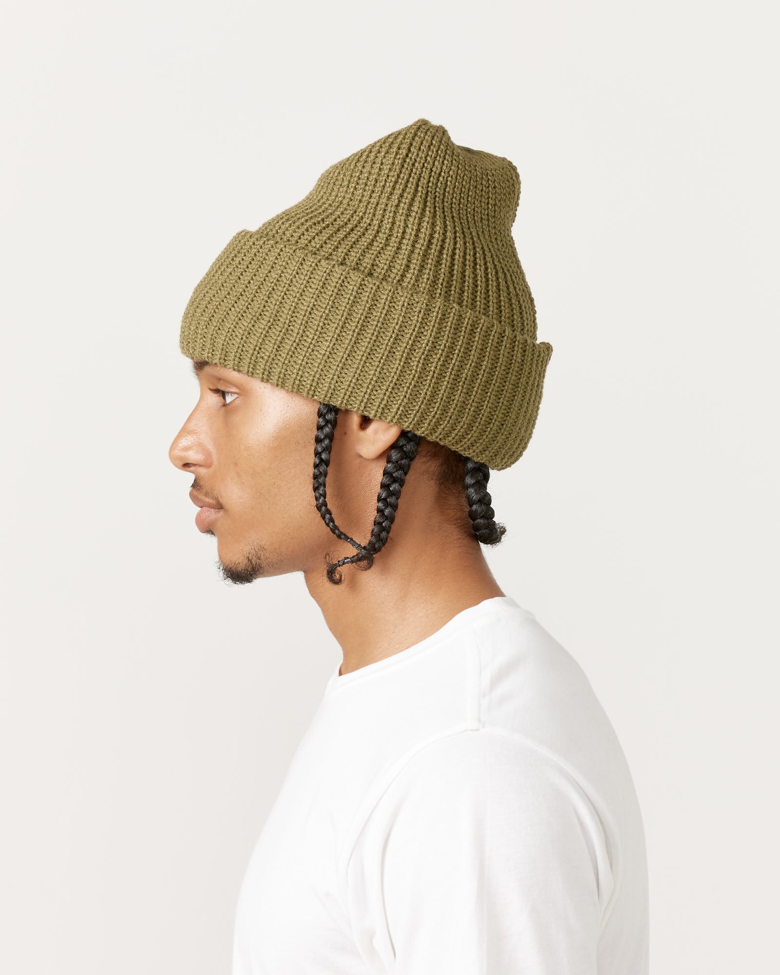 A man with two braids, shown in profile against a light background, wears the Rototo 5075 Chunky Beanie in Light Olive and a plain white shirt.