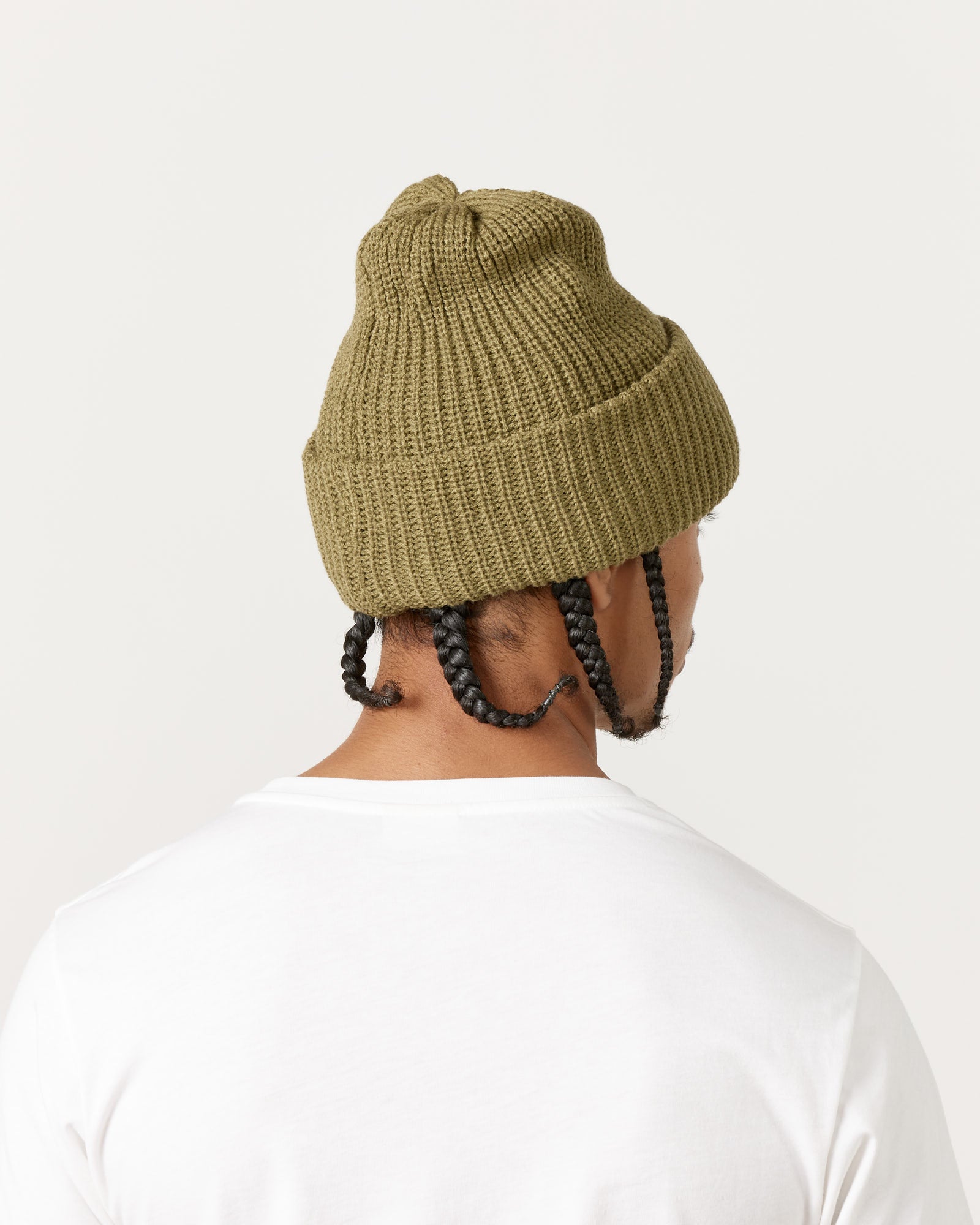 A person is seen from behind wearing a white shirt and the Rototo 5075 Chunky Beanie in Light Olive. Three braids fall from under the beanie, and the background is plain and light-colored.