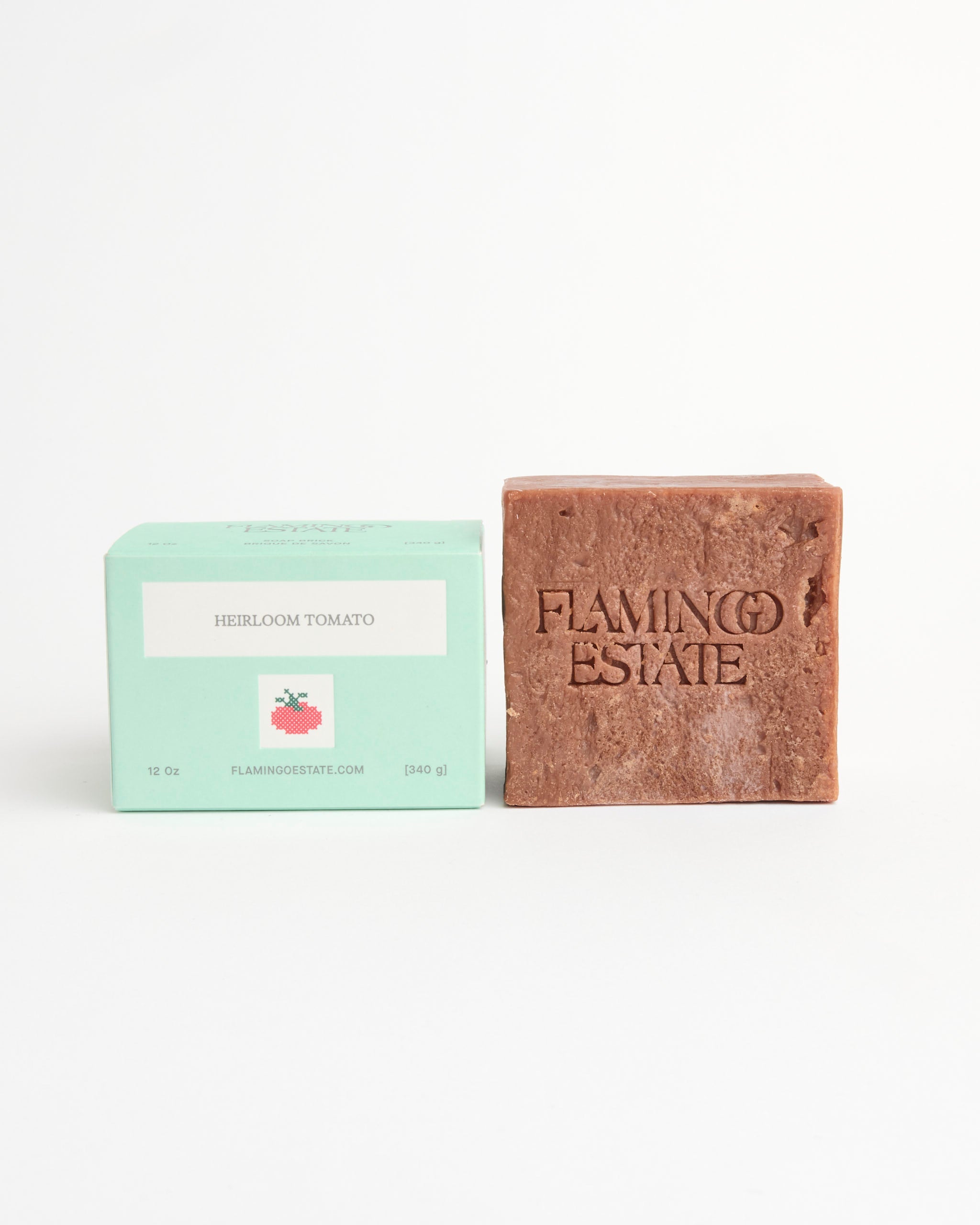 A brown handmade Heirloom Tomato Soap Brick by Flamingo Estate rests next to its green box with a tomato illustration, both displayed on a white background, highlighting the richness of plant oils.