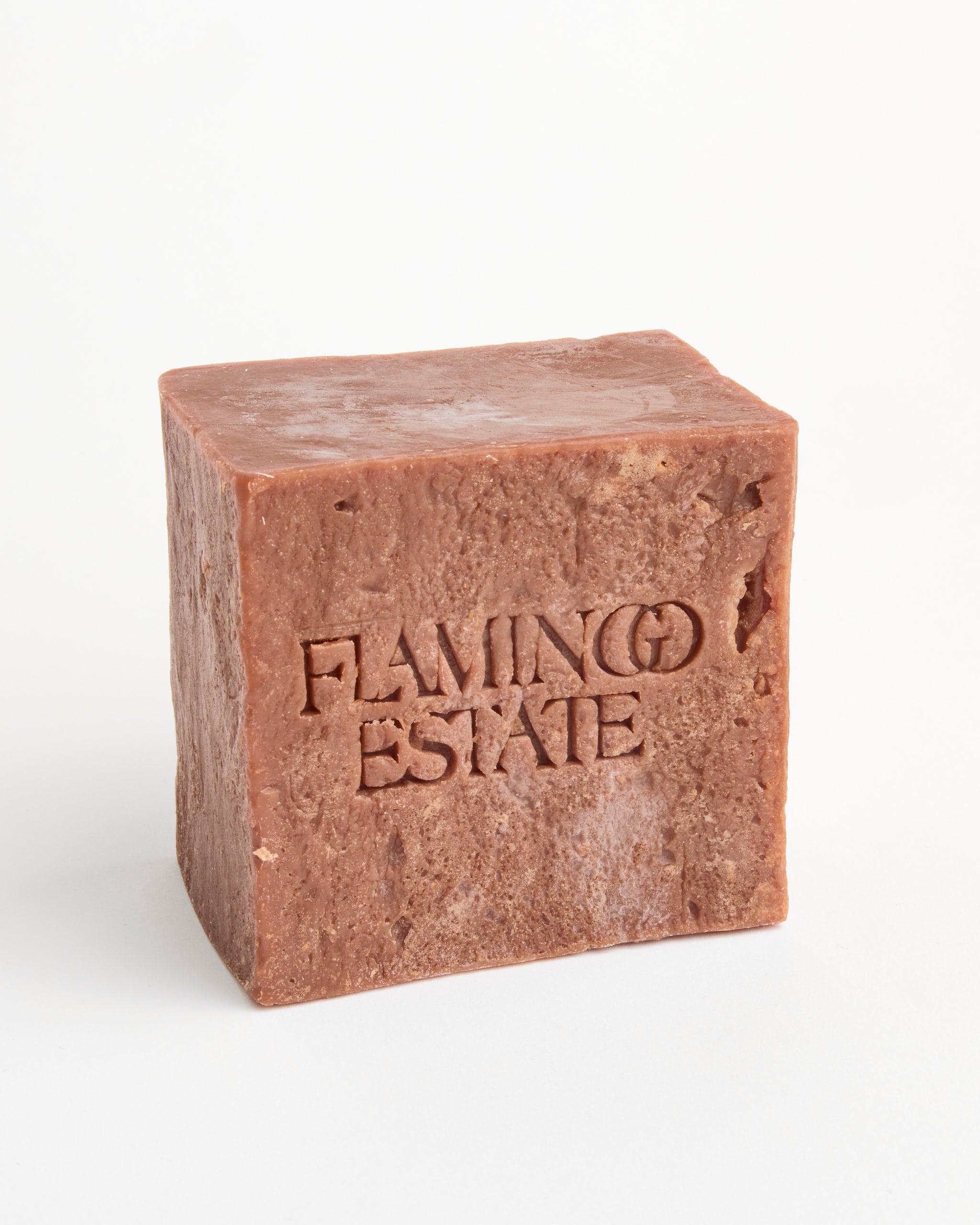 A handmade, rectangular Heirloom Tomato Soap Brick by Flamingo Estate, crafted from plant oils with rough edges and "FLAMINGO ESTATE" engraved on one side, appears reddish-brown against a plain white background.