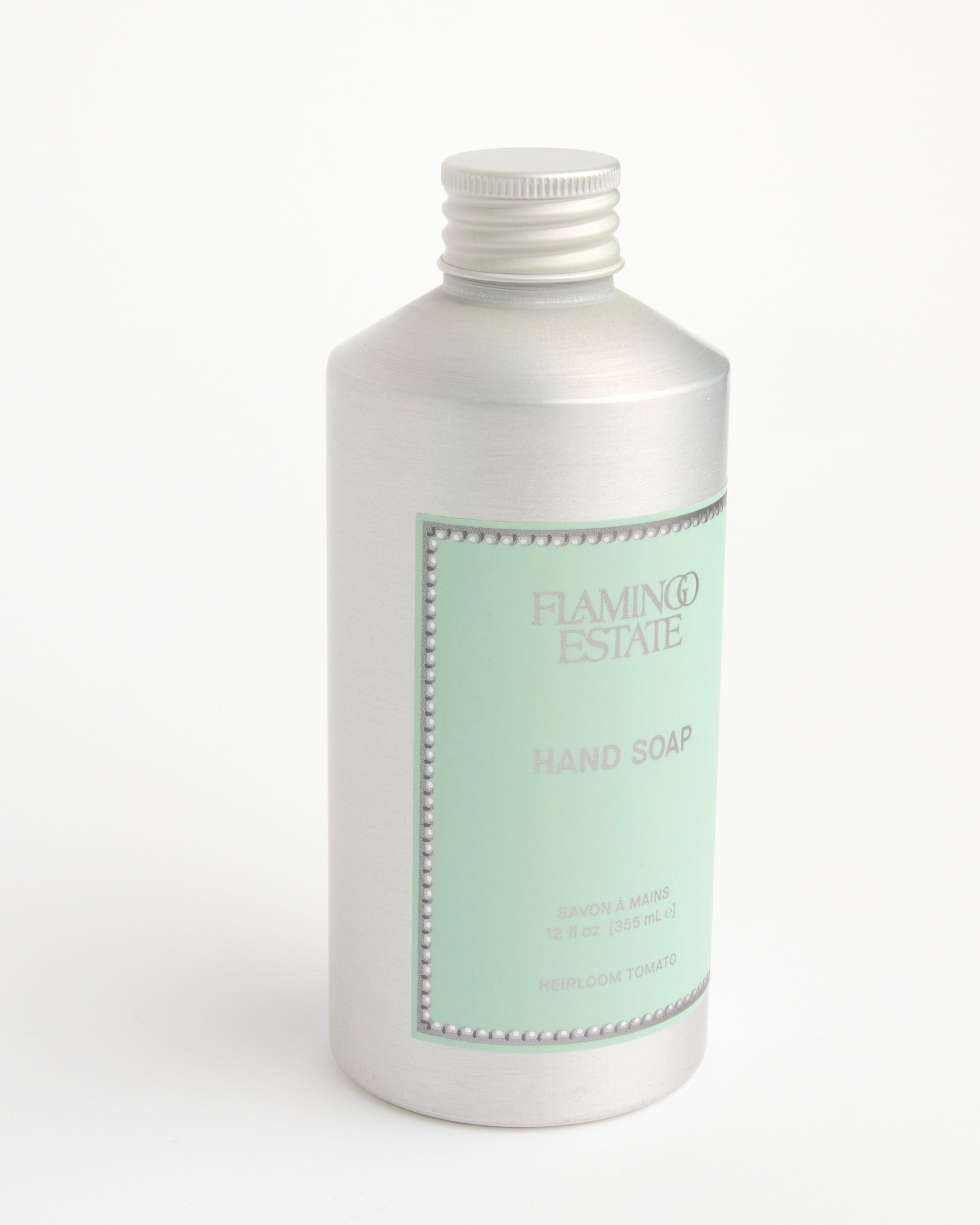 A silver bottle of Flamingo Estate Heirloom Tomato Hand Soap with a light green label, text, and beaded border sits uncapped on a white background.