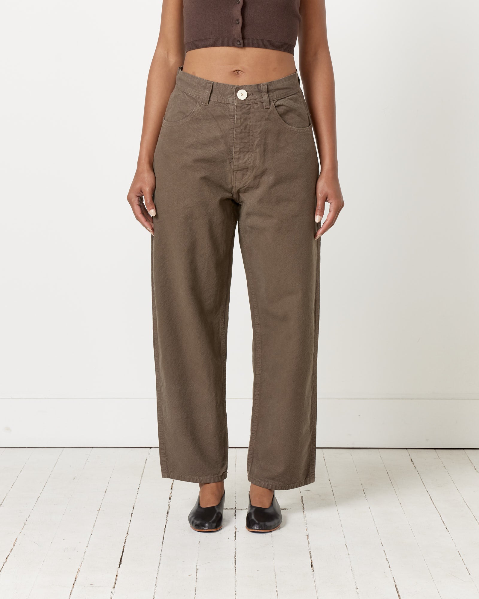 Someone stands on a white wooden floor against a plain wall, wearing Jesse Kamm’s California Wide Pant in Mushroom with a cropped brown top and black shoes. Their hands are relaxed by their sides.