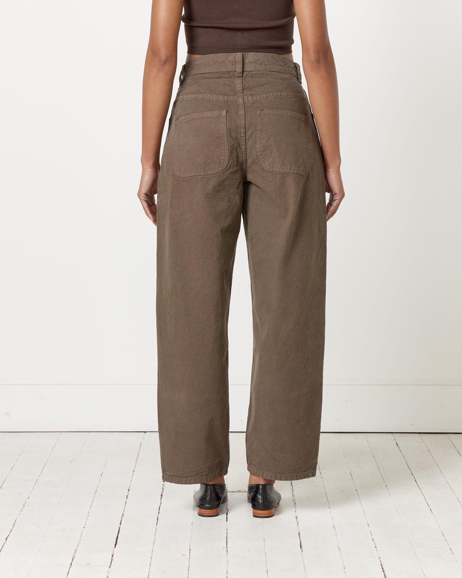 A person stands indoors facing away, wearing Jesse Kamm’s California Wide Pant in Mushroom, a dark brown sleeveless top, and black shoes. The setting is a white wood floor and plain white wall. Made in California.