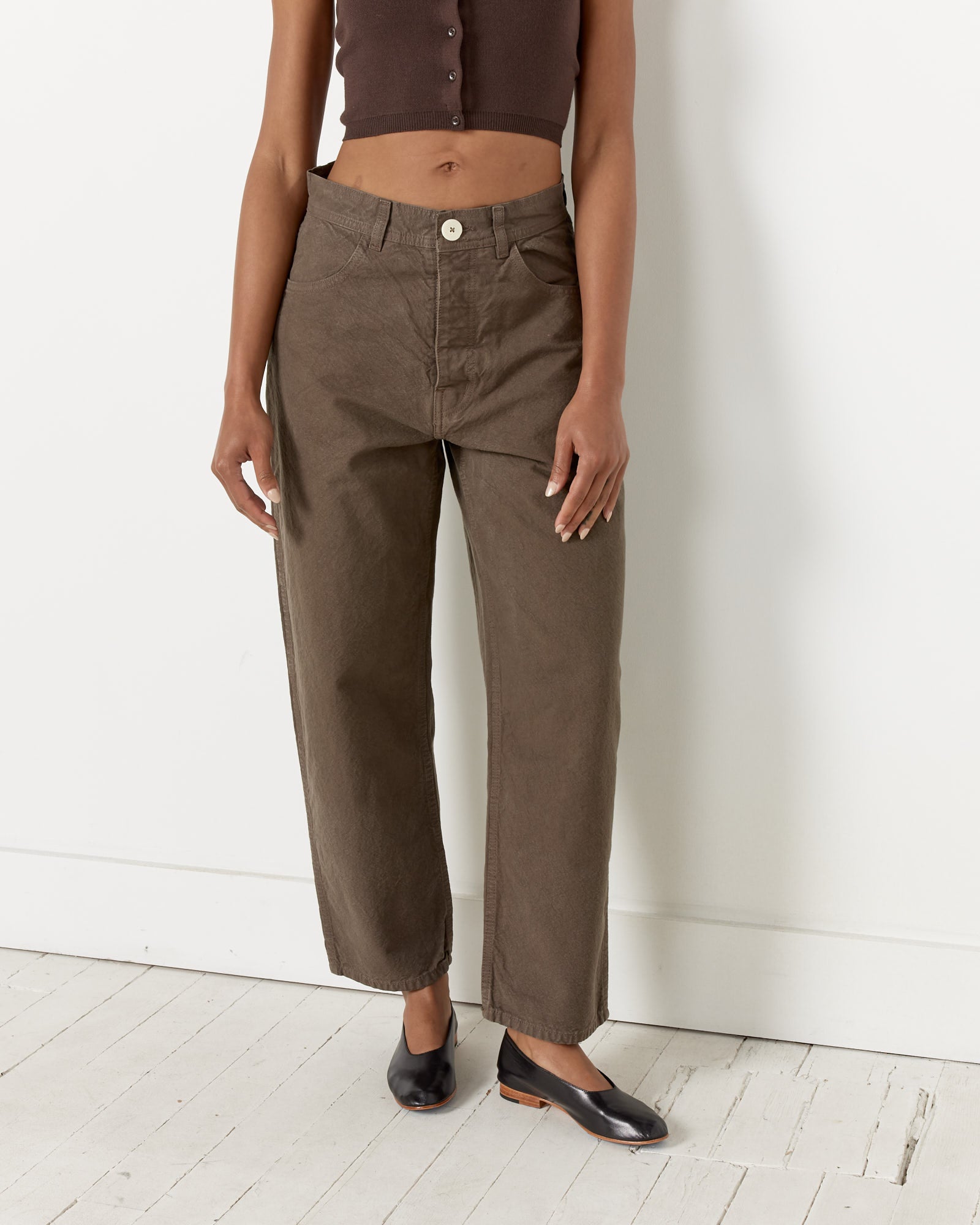 Wearing Jesse Kamm's California Wide Pant in Mushroom, a dark sleeveless top, and black slip-on shoes, a person stands against a white wall on a light wooden floor with their head out of frame.