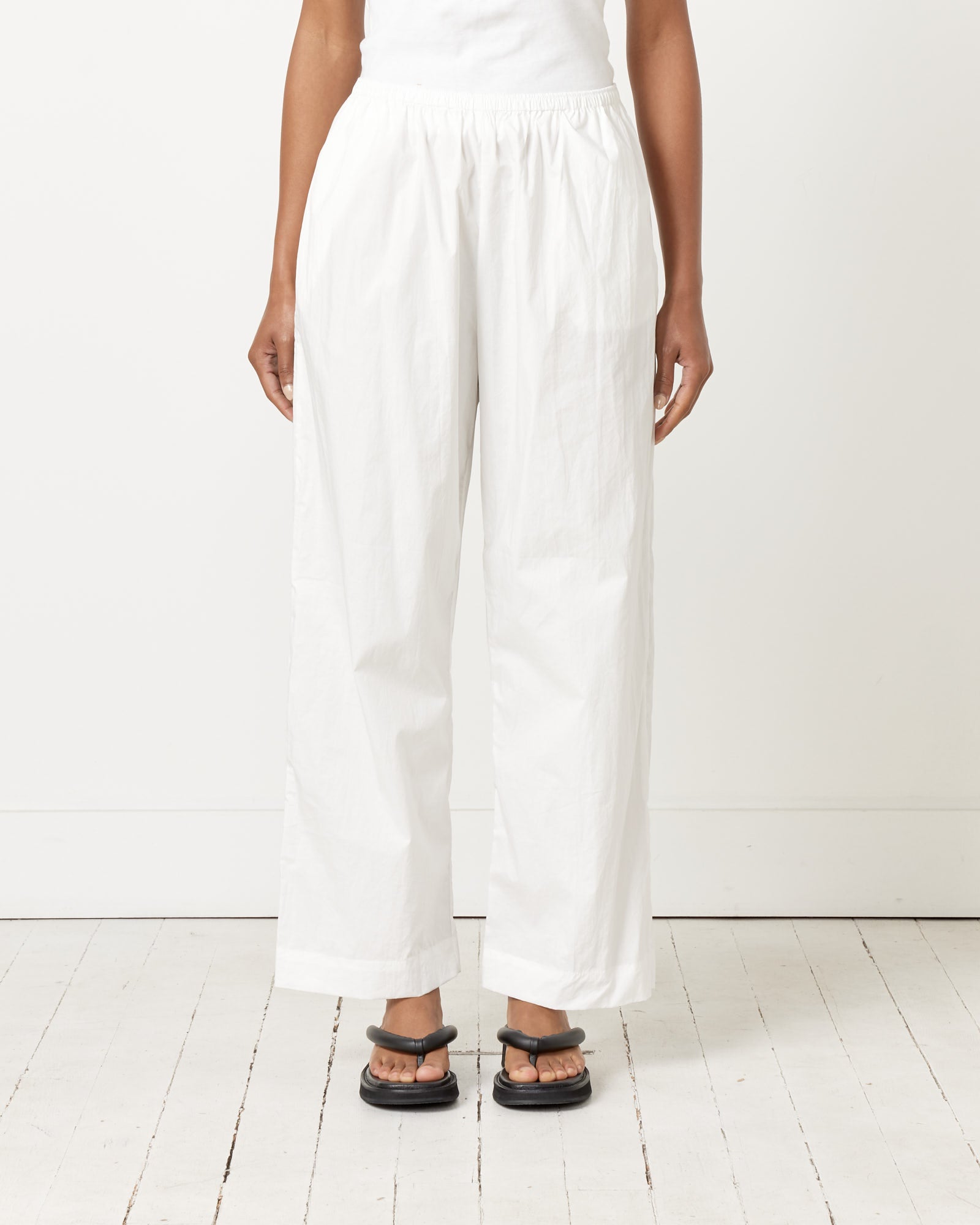 A person wearing Deiji Studios’ Ease Trousers in White and black sandals stands on a white wooden floor against a plain background. Only the lower body is visible, emphasizing the relaxed style of the Ease Trousers.