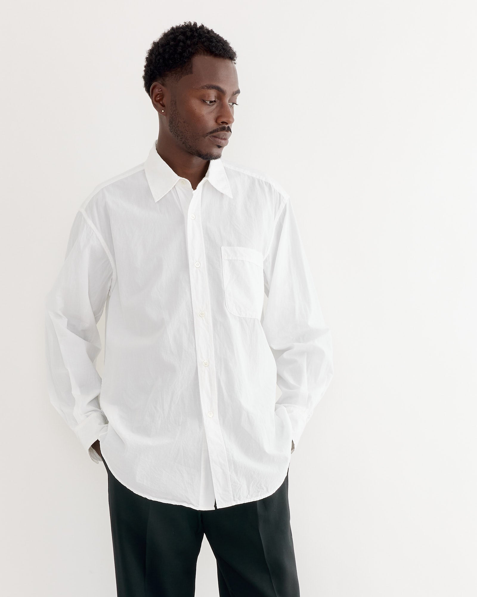 A man wears the SMOCK Gio Shirt in Crushed Cotton White with black pants, standing against a plain white background and looking to his left with hands in his pockets.