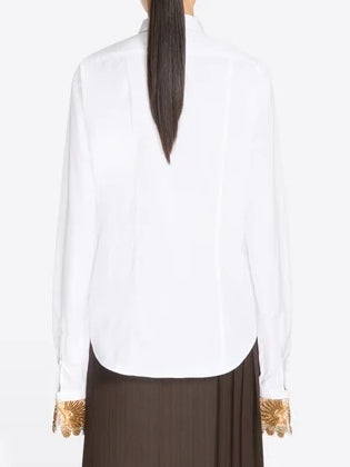 A person with dark hair pulled back is seen from behind, wearing the Dries Van Noten Embellished Shirt in White— a long-sleeved cotton top featuring gold ornate, embellished cuffs—paired with a dark brown pleated skirt.