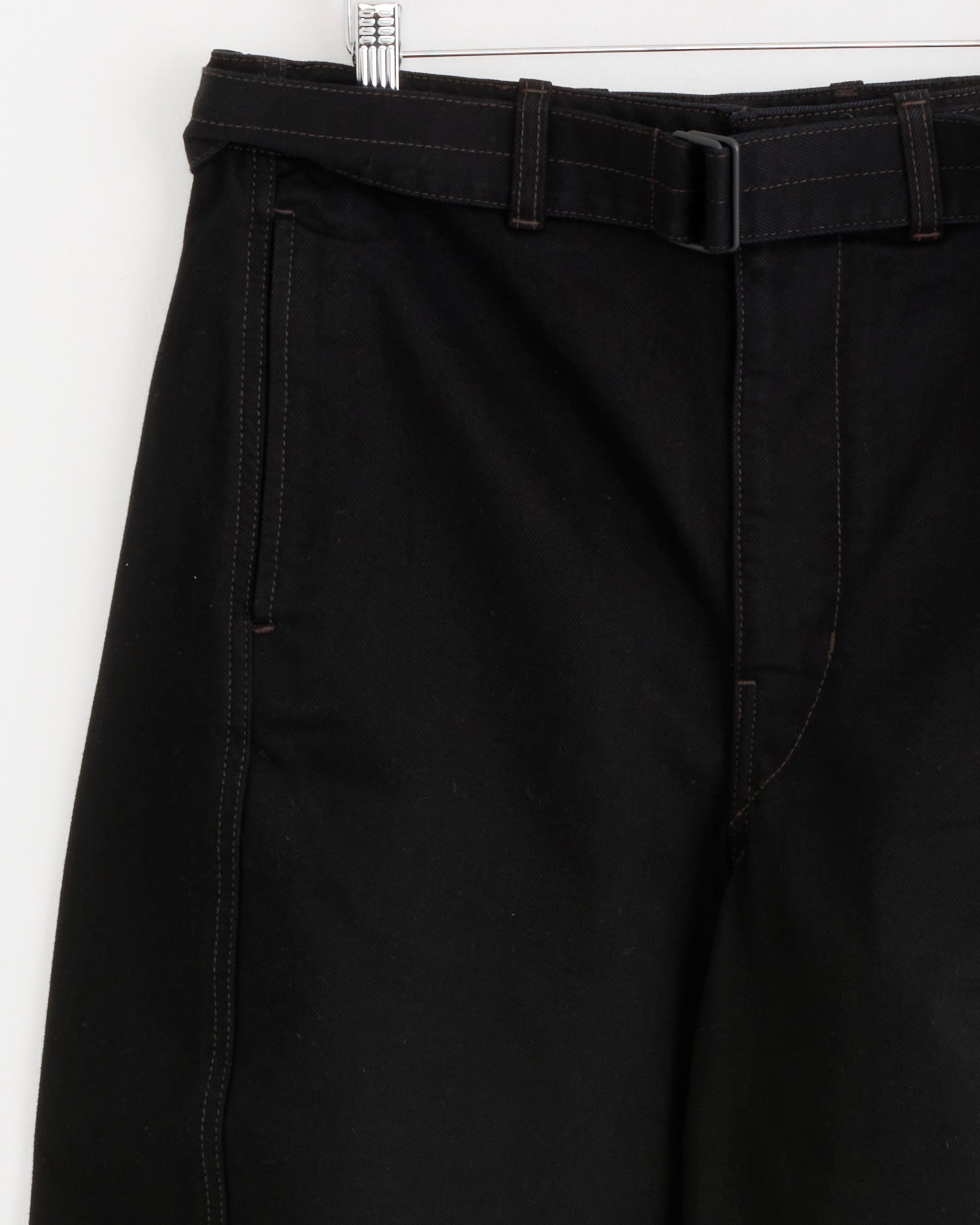 Close-up of the Lemaire Twisted Belted Pant in Black hanging, showcasing a black belt with a rectangular buckle, dark brown stitching, and prominent seams along the pocket and leg. Made from heavy denim, these unisex pants are displayed with only the upper portion visible.