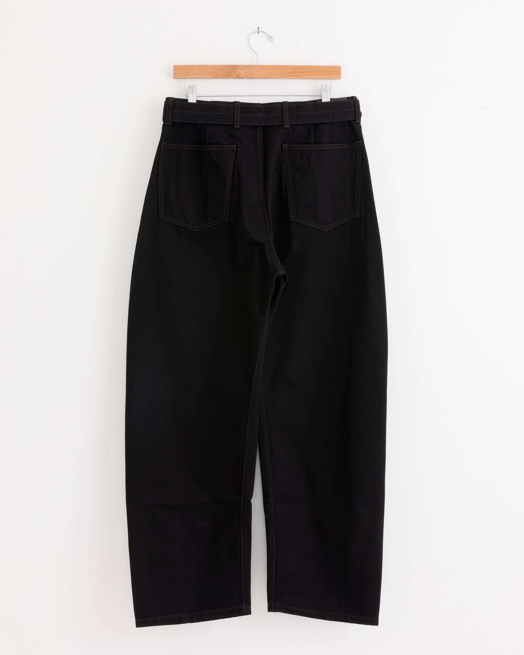A pair of Lemaire Twisted Belted Pant in Black, unisex jeans featuring belt loops and two back pockets, crafted from heavy denim, displayed on a wooden hanger against a white background.