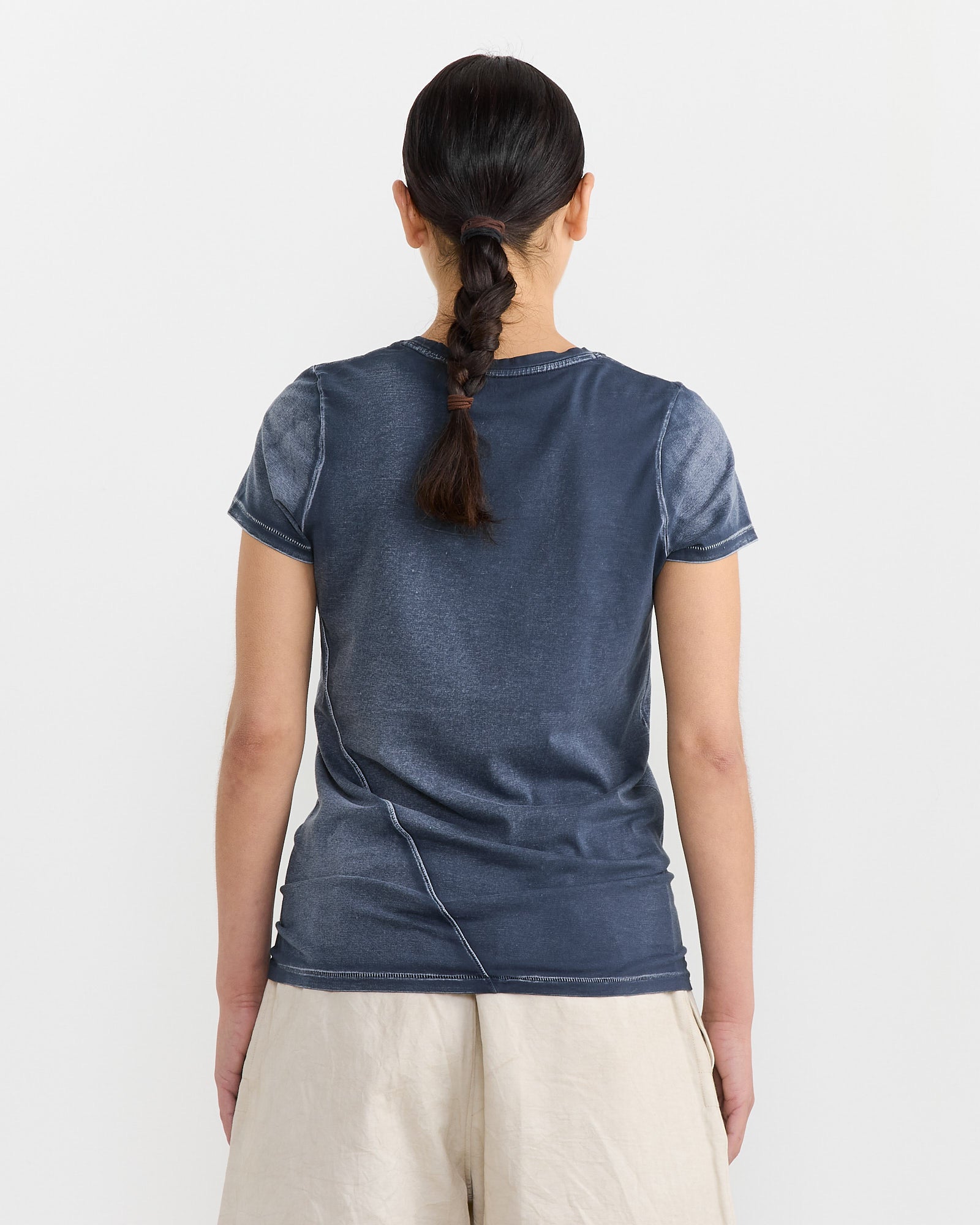 A person with dark braided hair stands with their back to the camera, wearing a Bias Baby Tee in Indigo by Lauren Manoogian and light beige pants. The Pima cotton shirt adds a soft touch against the plain, light-colored background.