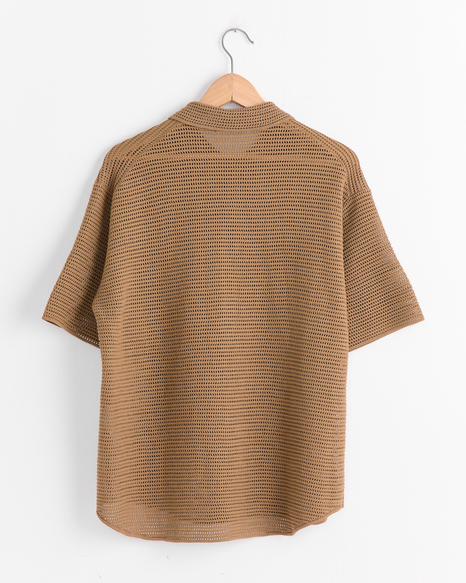 The Auralee Mesh Knit Half Sleeved Shirt in Khaki Brown hangs on a wooden hanger against a white wall. The relaxed-fit shirt, made of cotton yarn, showcases a crochet-like texture with its loose pattern, displayed back-facing forward.