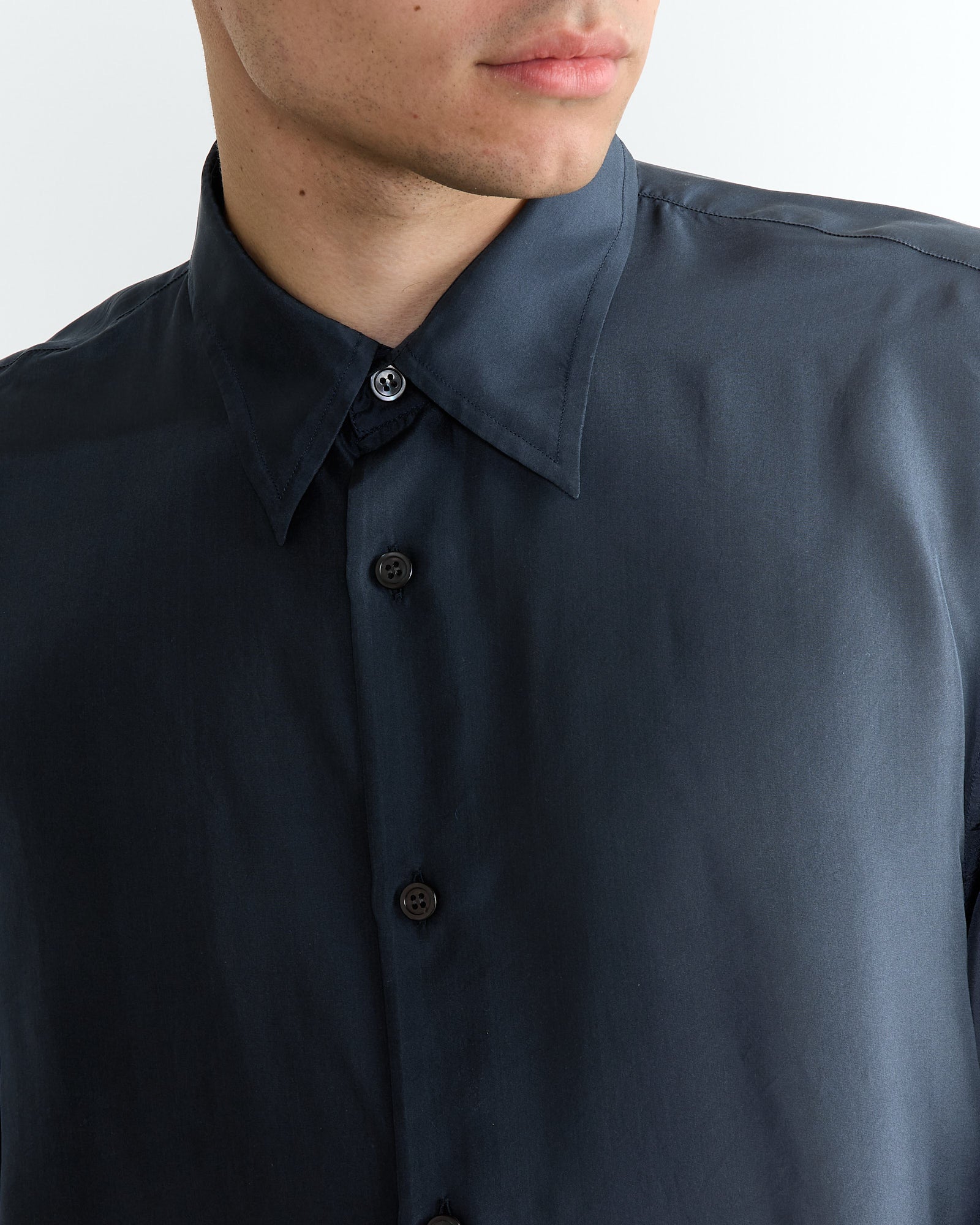 A person wearing a navy, long-sleeve Dries Van Noten shirt with a classic collar. The silk fabric has a smooth texture, and the person is slightly angled to the right, revealing just part of their face and upper body.