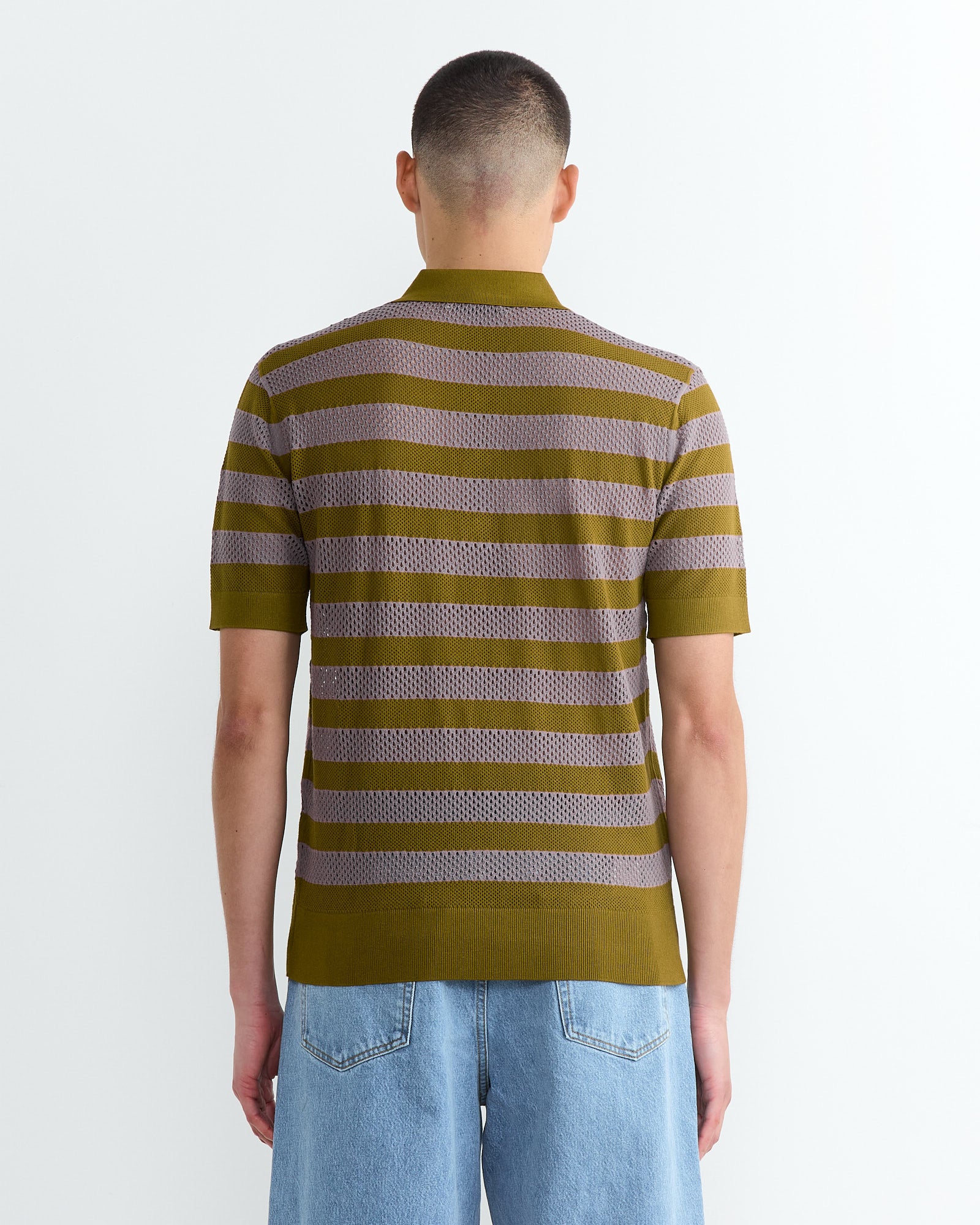 A person with short hair, viewed from behind, wears a Dries Van Noten Stripe Knit Polo in olive and brown stripes with light blue jeans. The knit polo stands out against the plain white wall backdrop.