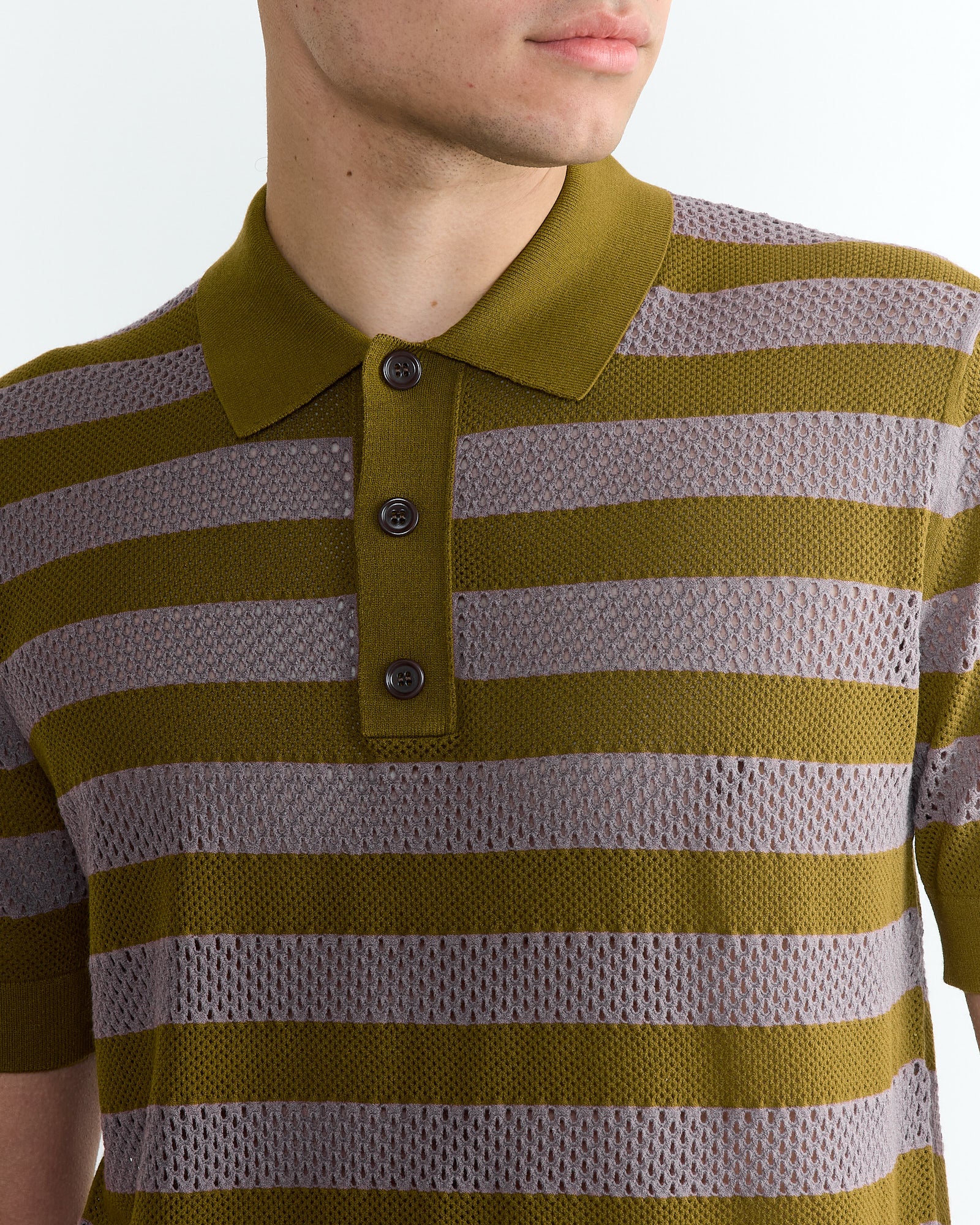 A person wears the Stripe Knit Polo in Olive by Dries Van Noten, featuring olive green and gray horizontal stripes, a collar, and three buttons. The background is plain white.