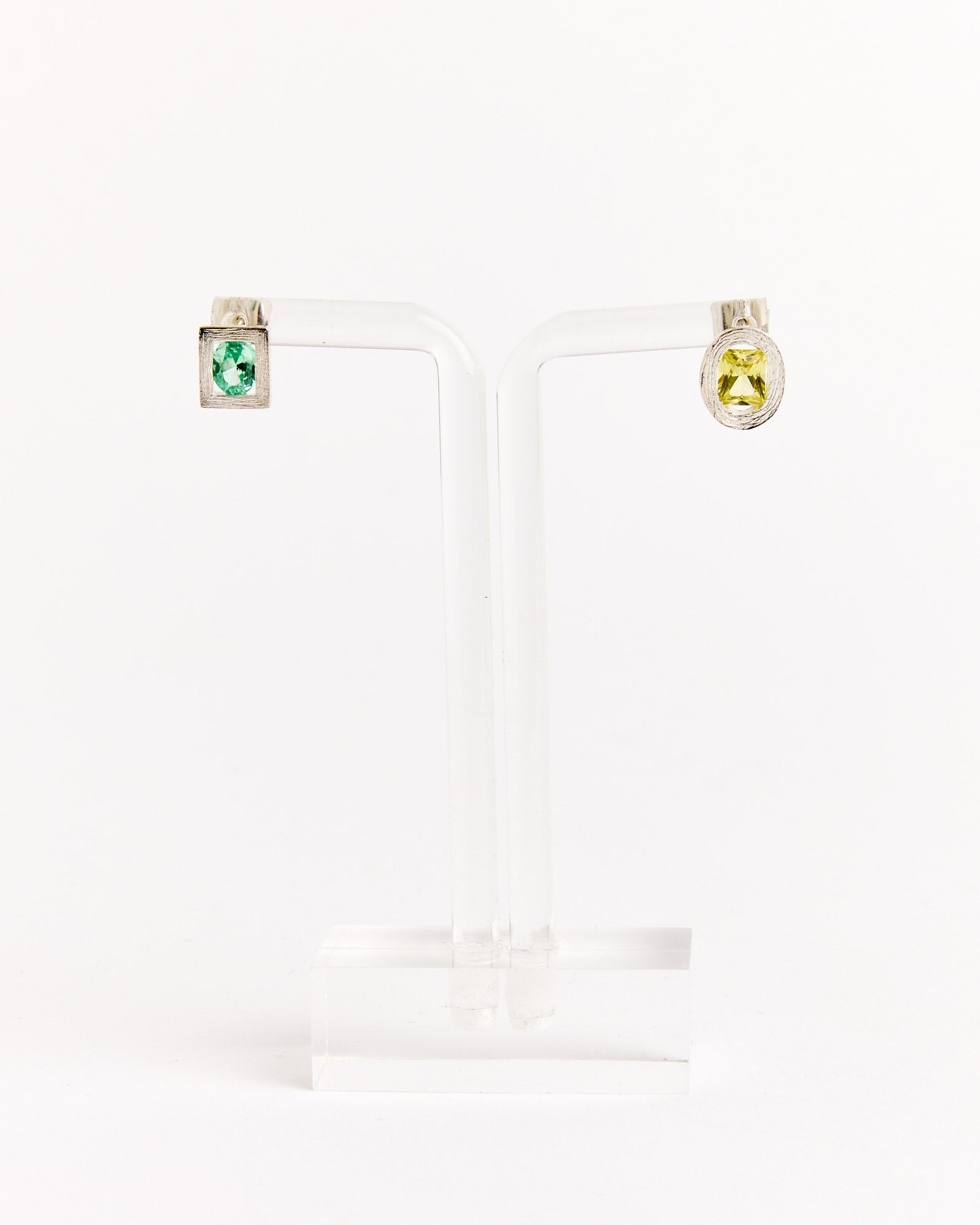 Bleue Burnham’s Opposite Earrings in Silver/Green/Yellow are mismatched studs—one with a green square and the other with a yellow oval gemstone—displayed on a clear stand against a plain white background.