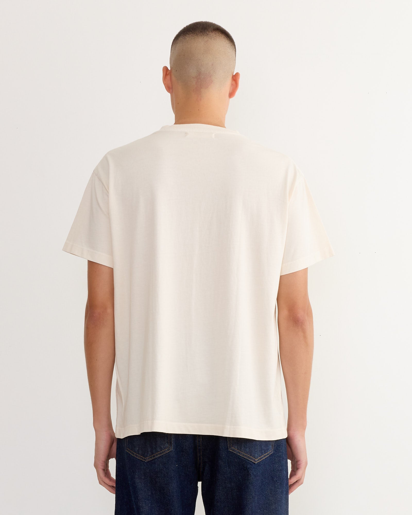 A person with short hair stands facing away, wearing the SMOCK Big Pocket Tee 3 Pack in Natural and dark blue jeans against a plain white background.