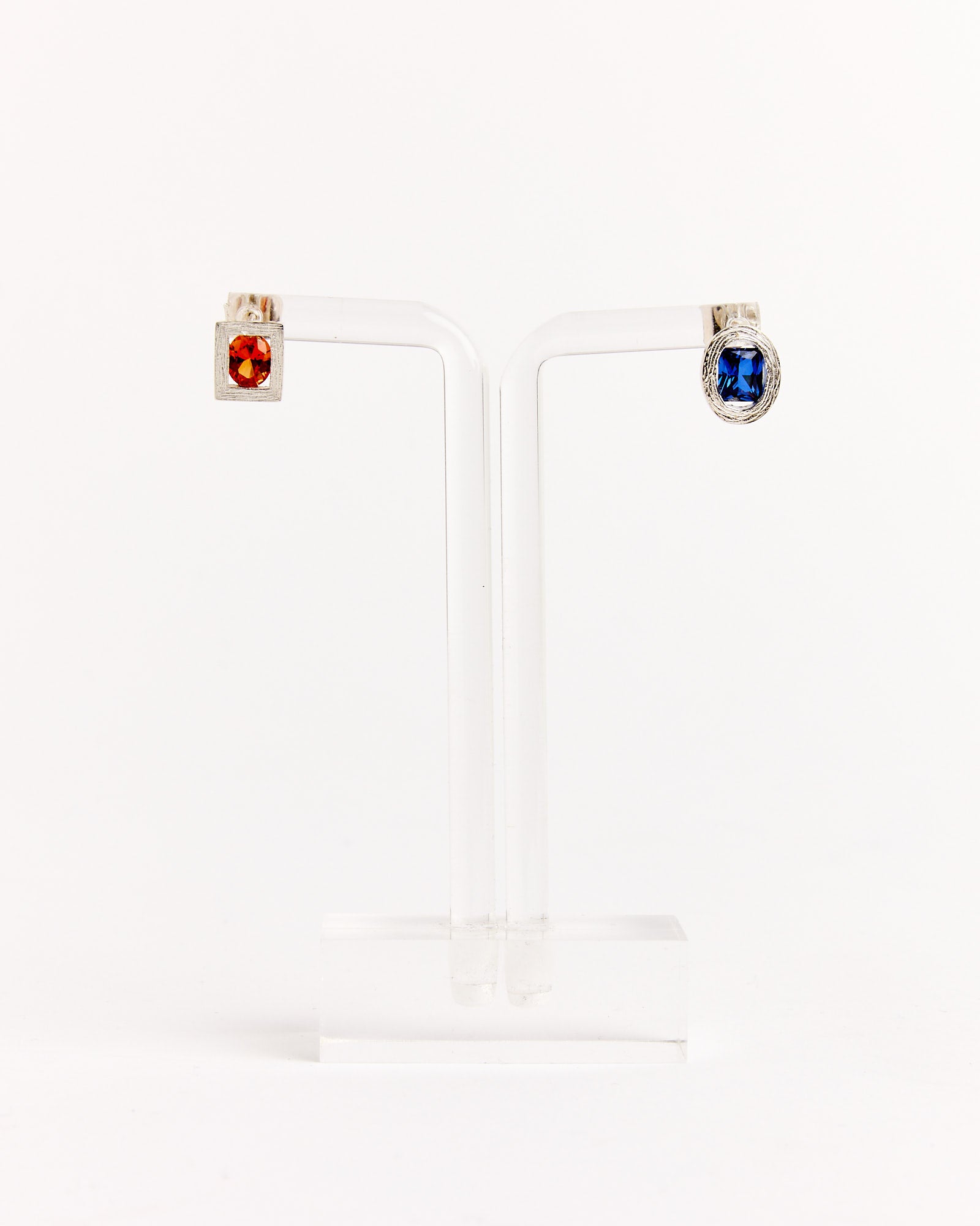The Bleue Burnham Opposite Earrings in Silver/Orange/Blue feature mismatched studs on a clear stand—one with a square orange gemstone, the other with a round blue gemstone—both set in silver-toned metal.