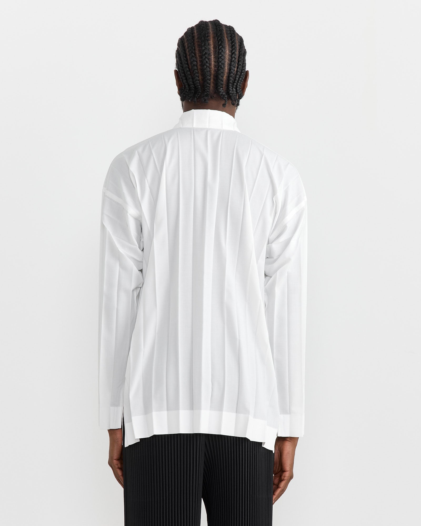 A person with braided hair is seen from the back wearing a long-sleeved Edge Shirt in White by Homme Plissé Issey Miyake, featuring signature pleating, paired with black pleated pants against a plain white background.