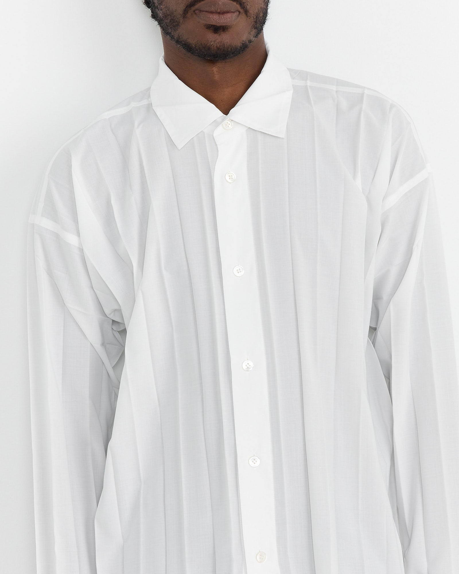 Against a plain white backdrop, someone dons a loose-fitting Homme Plissé Issey Miyake Edge Shirt in White with signature pleating and subtle vertical stripes. The shirt features a classic collar and long sleeves while the persons face is partially out of the frame.