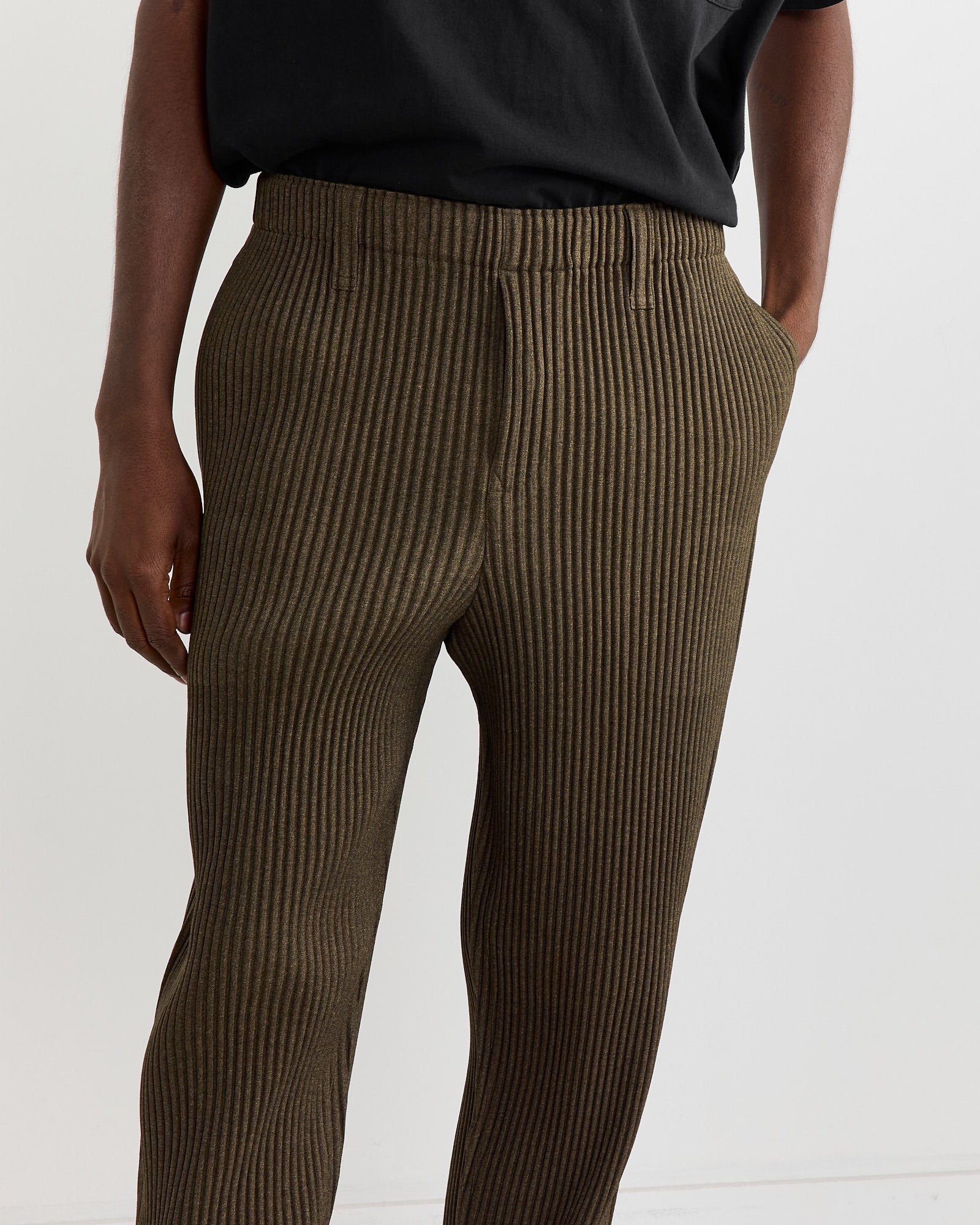 A person is wearing the Wool Like Light Pant in Khaki by Homme Plissé Issey Miyake, featuring a fitted waist and straight legs. Paired with a black shirt, hands in pockets, this stylish ensemble channels effortless elegance.