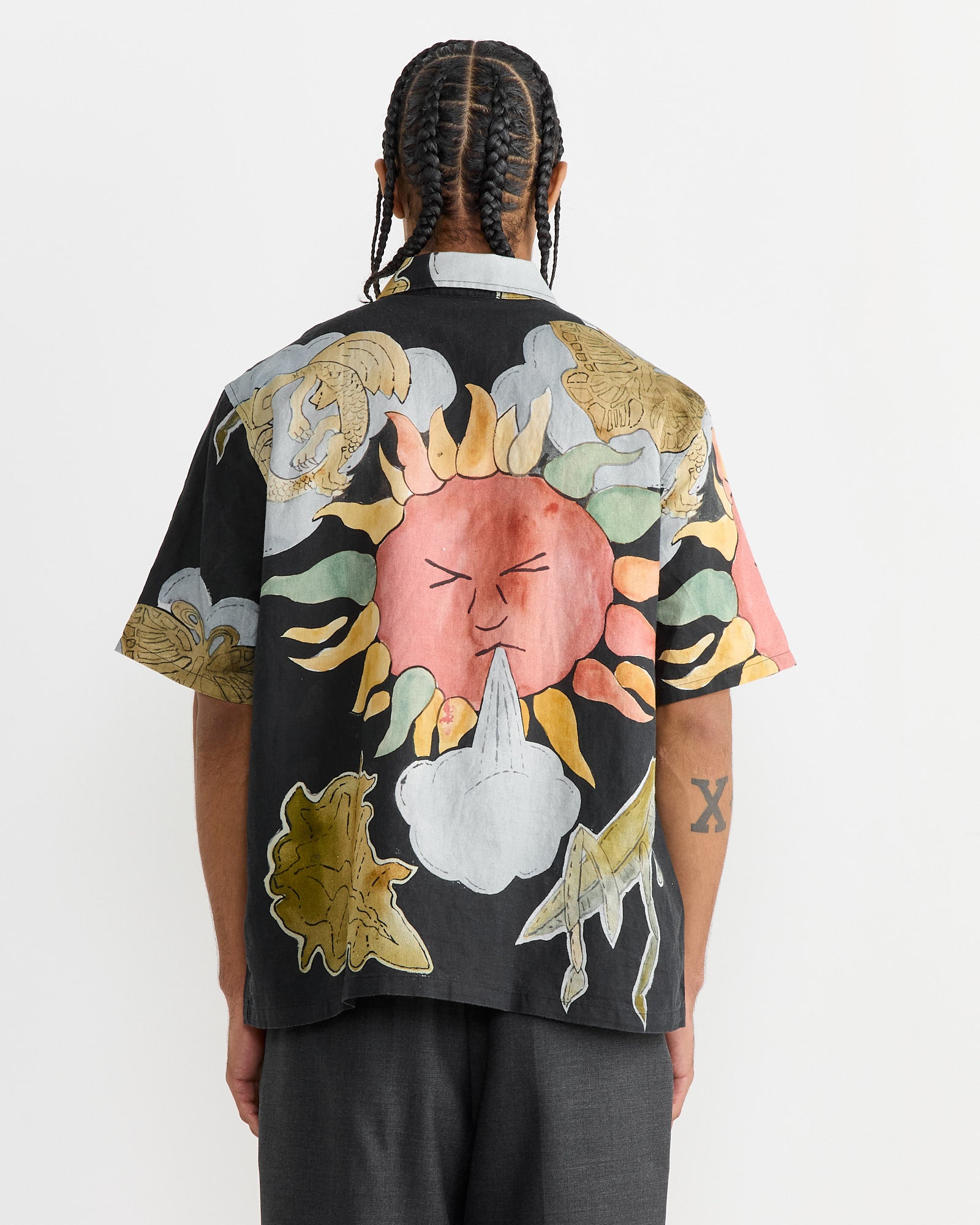 A person with braided hair faces away, wearing the “Box Shirt in Solar Power Print” by Our Legacy. The black short-sleeve shirts boxy fit boasts a vibrant array of abstract designs—sun-like faces, animals, and clouds. Gray pants complete the look as they reveal a forearm tattoo against a plain white backdrop.
